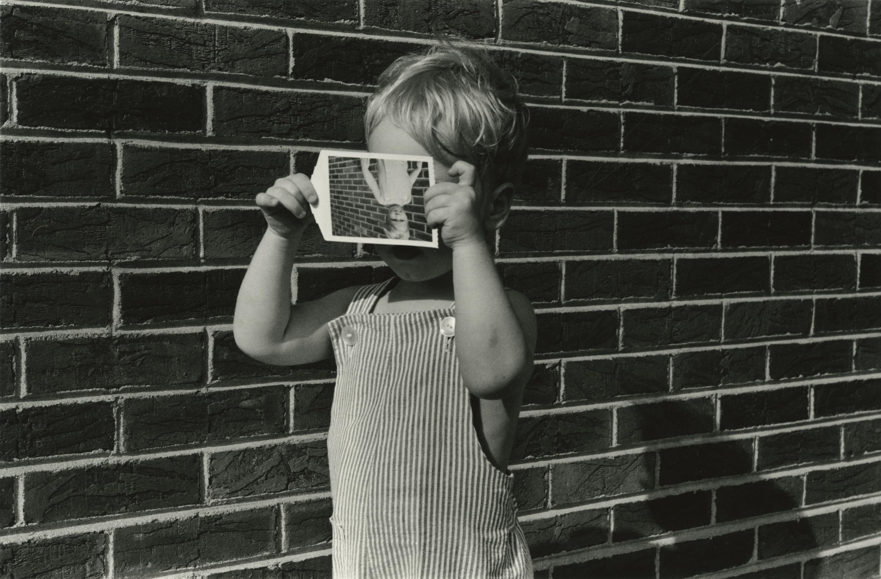 A child stands in front of a brick wall, holding in front of his face an upside-down snapshot of himself standing in front of a brick wall.