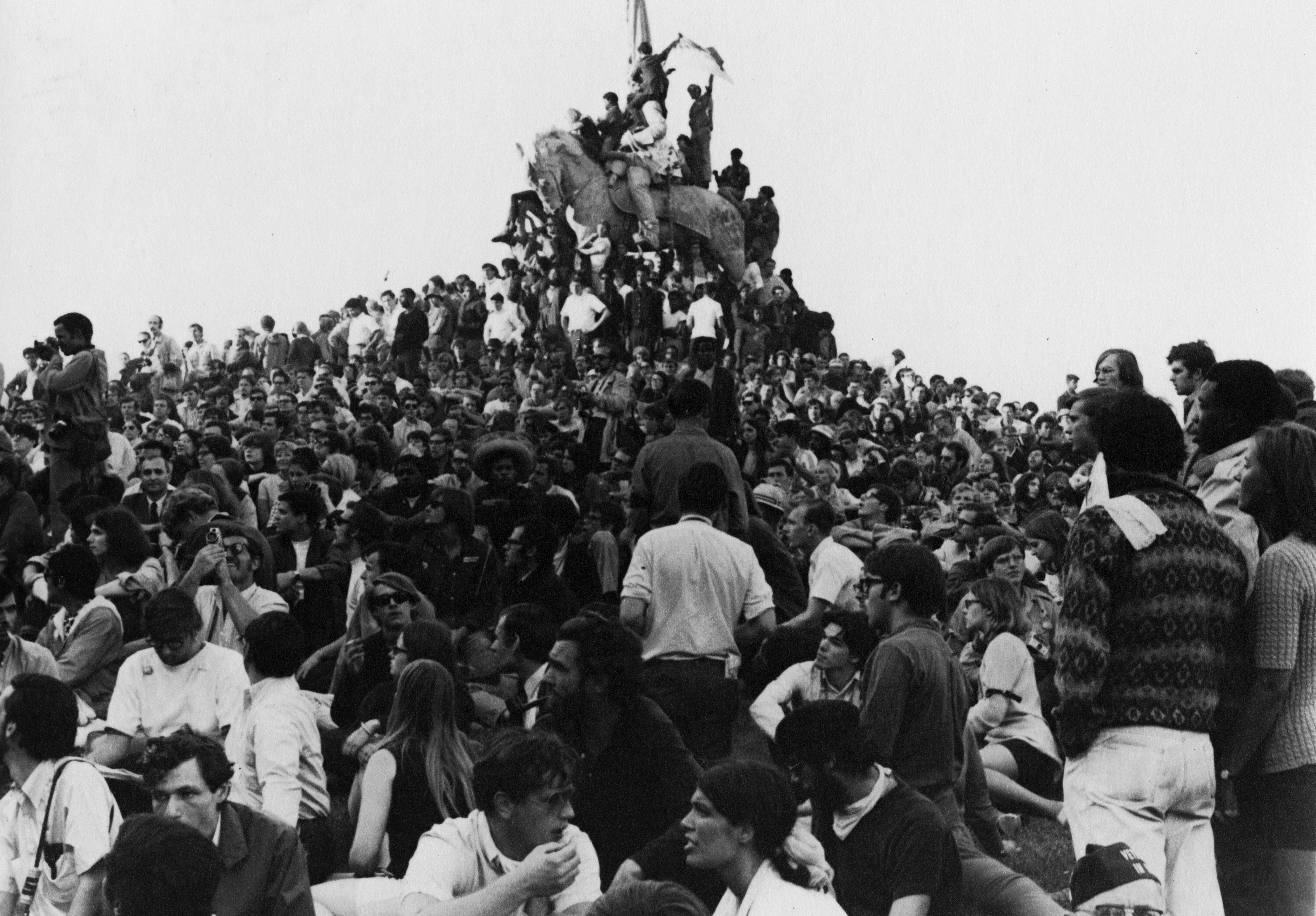 A black-and-white image shows a dense crowd of diverse young people gathered around and upon an elevated statue of a horse. Some people are climbing the horse, with the person at the top waving a flag.