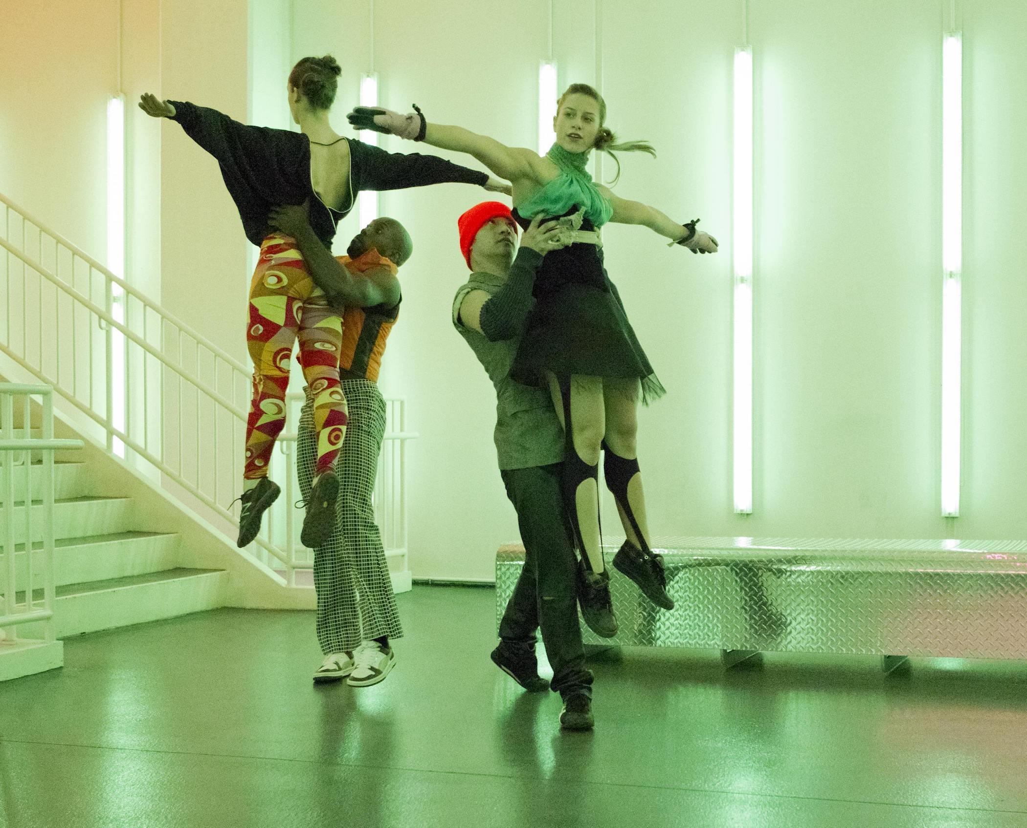 Two make dancers hold aloft two female dancers in front of a stairwell in a room lit with vertically installed neon tube lights.
