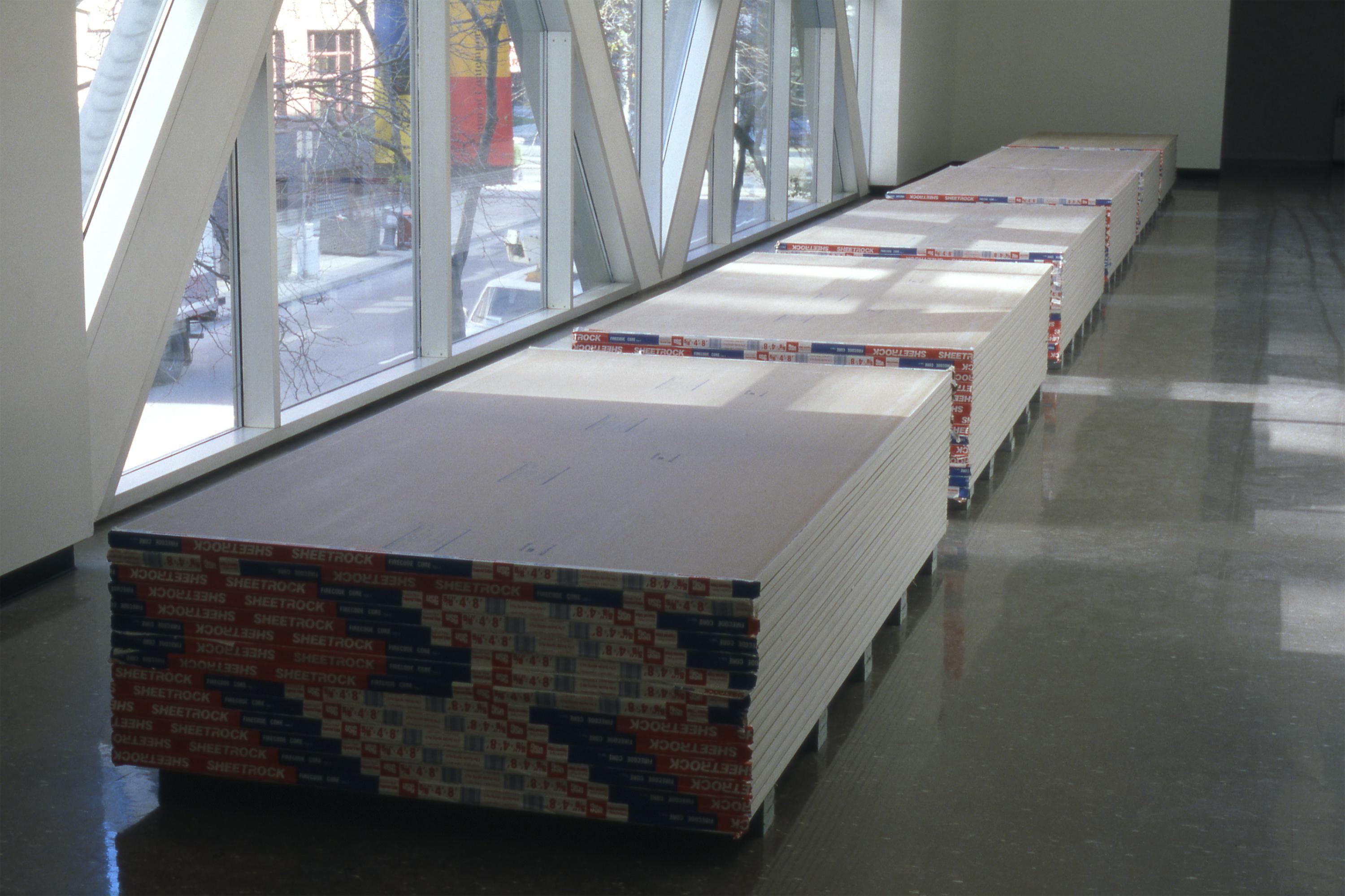 Gallery view of four equal stacks of drywall in front of a window.