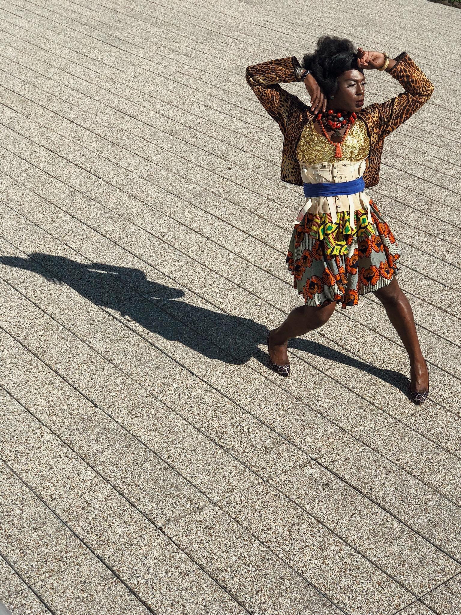 A dark complected person wearing a jacket, corset and many necklaces over an African print dress stands with splayed legs and upraised arms in a bright outdoor space.