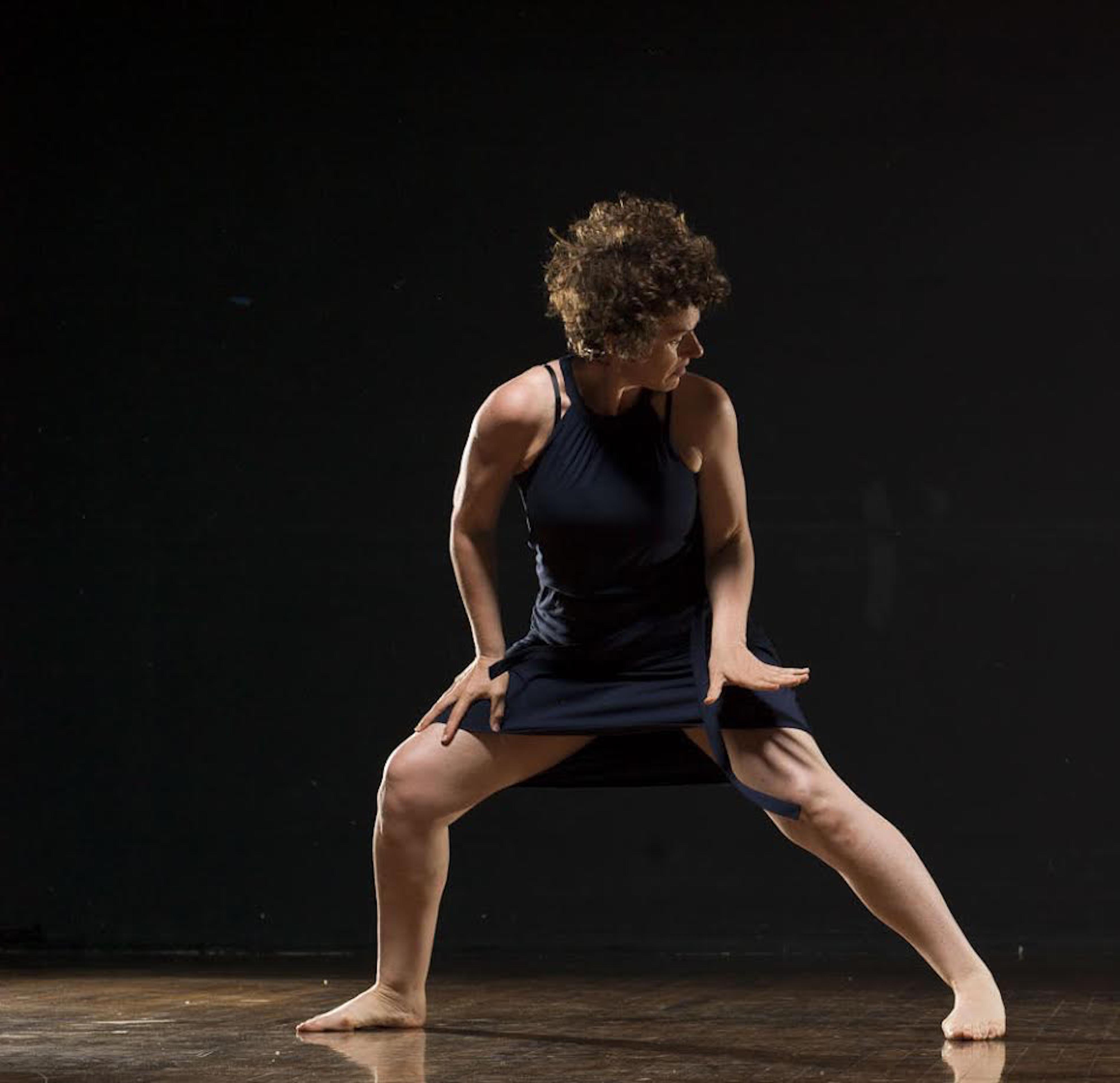 A barefoot dancer in a black dress lunges in a spotlight against a dark background.