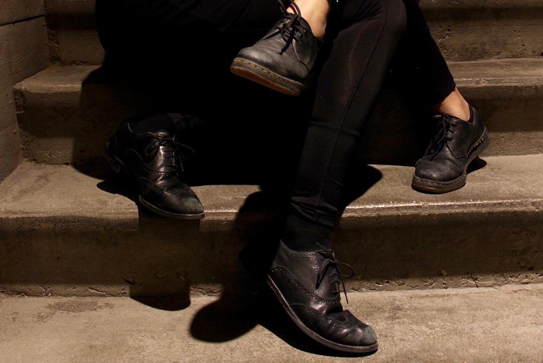 Two pairs of legs in black garments and black shoes are intertwined, seated on concrete steps