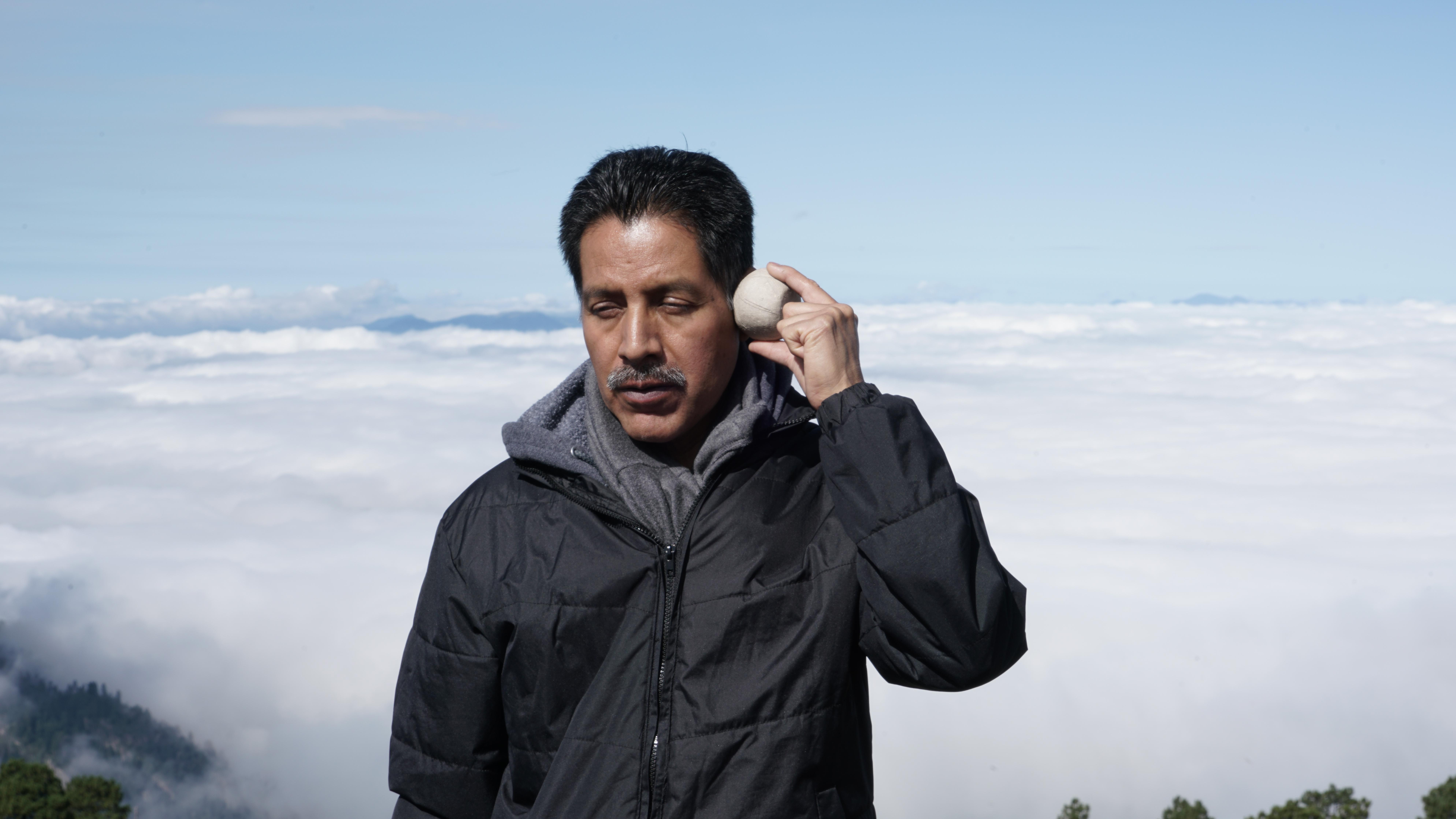 A man with medium-toned skin closes his eyes and holds a round, rock-like object against his temple. Behind him is a low cloud bank, indicating that he is standing at a very high elevation.