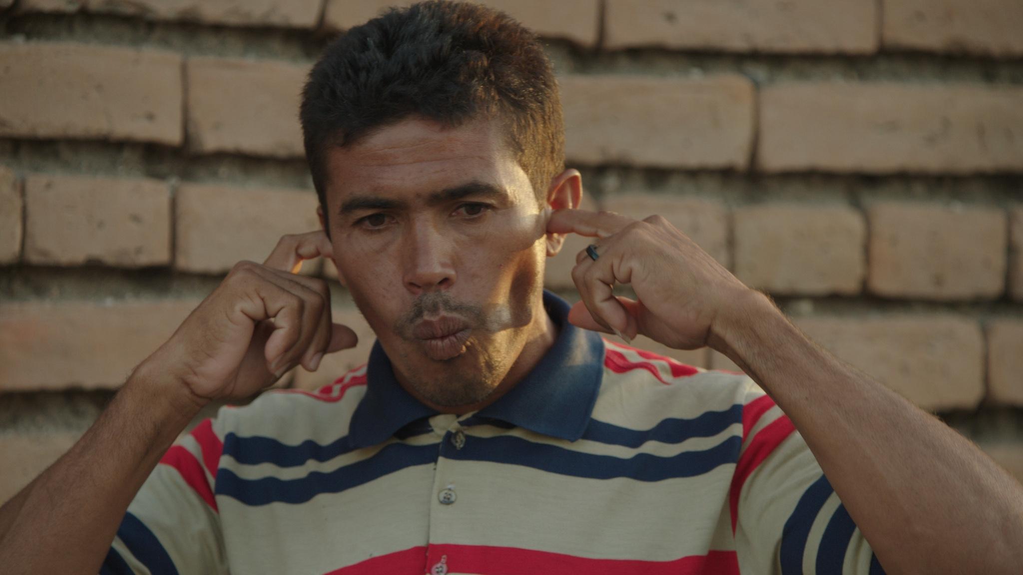 A man wearing a striped polo shirt purses his lips while placing his index fingers in each ear.