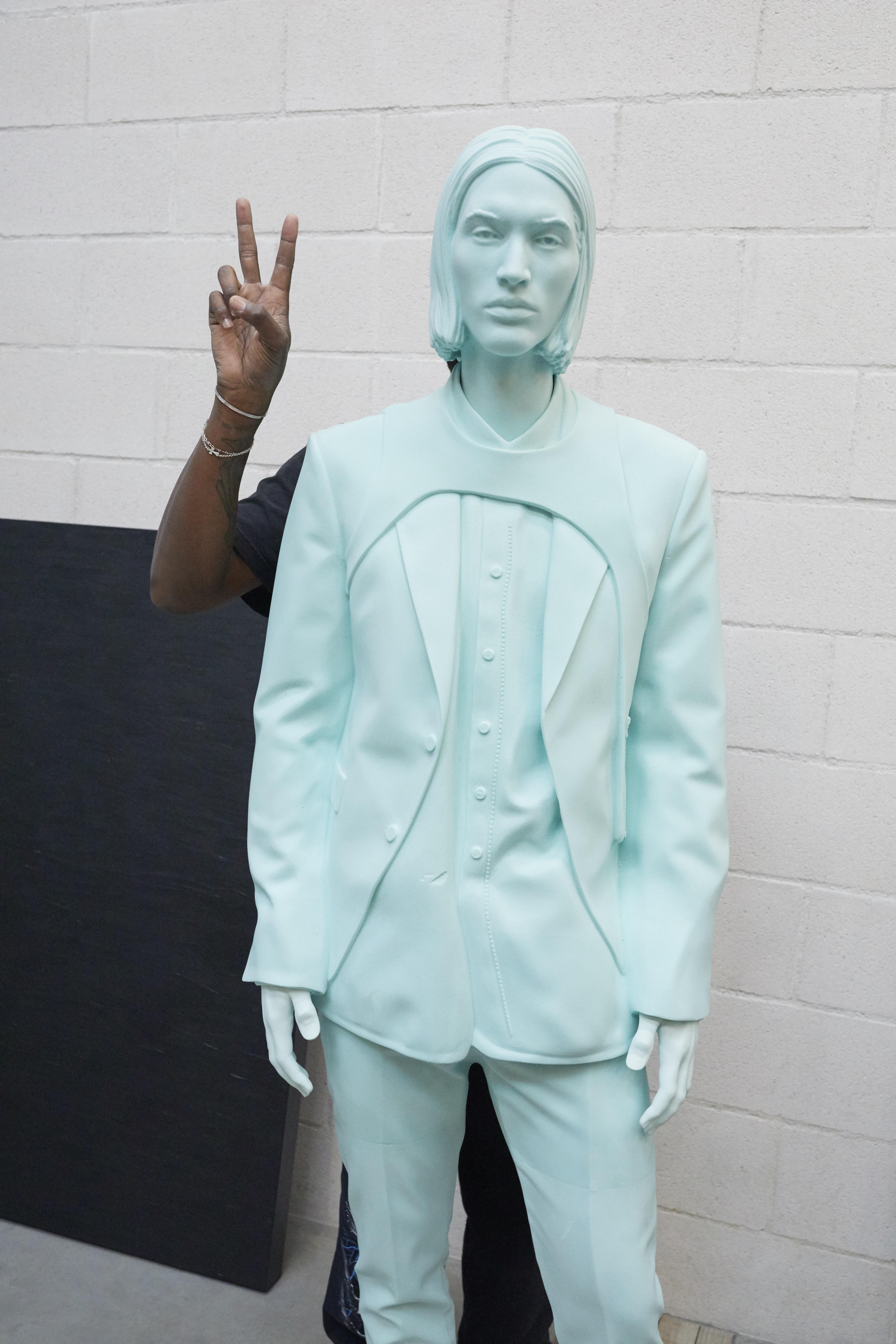 A dark-skinned arm holds up two fingers in a peace symbol from behind a monochrome light-blue mannequin.