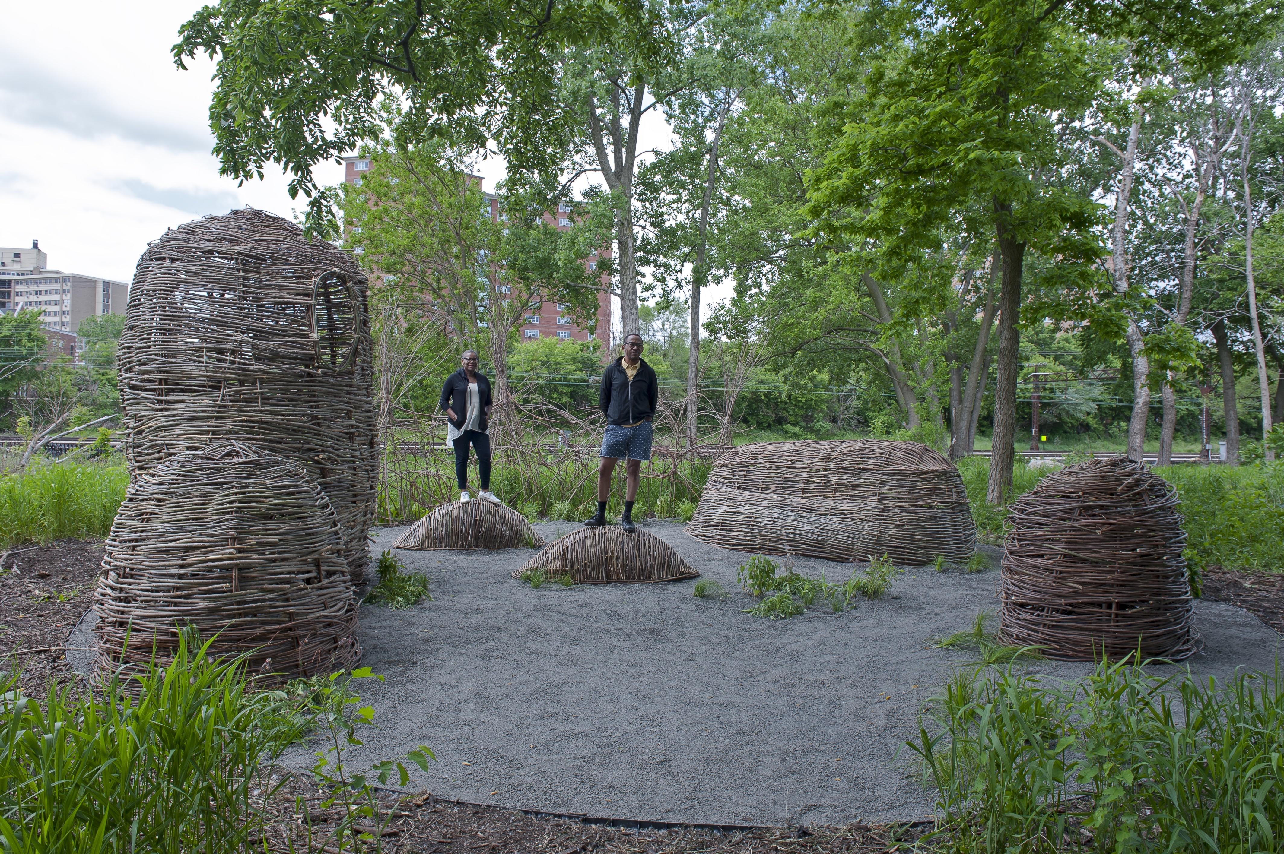 Two people stand on small mounds that are woven like wicker baskets. They are surrounded by four other similar mounds and urban greenery.