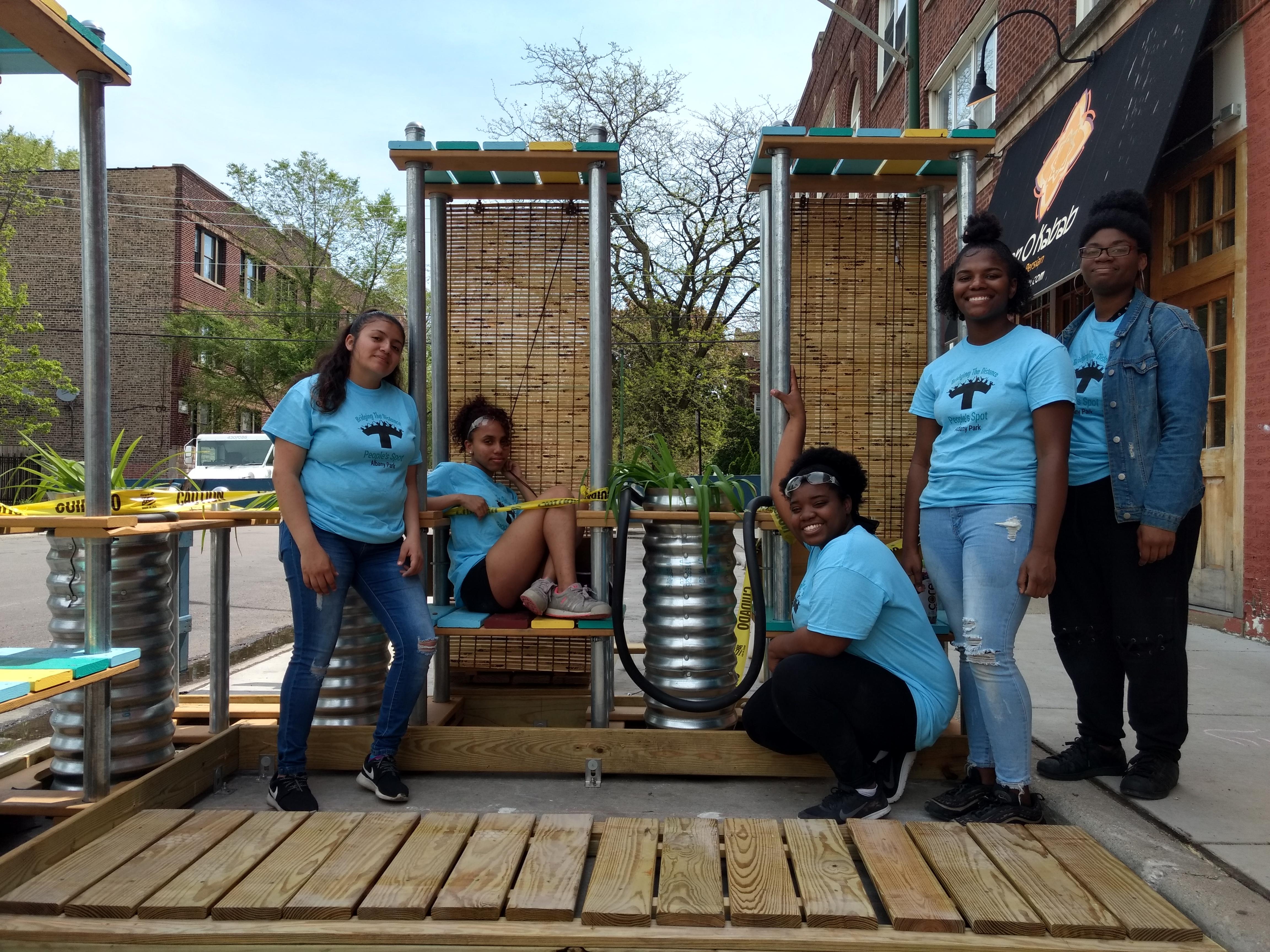 A group of five young people pose in front of a structure made of wood, metal, and rattan.