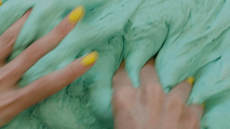 In a short animation, light-skinned hands with vivid yellow finger nails repeatedly massage a large mass of mint-green goo.