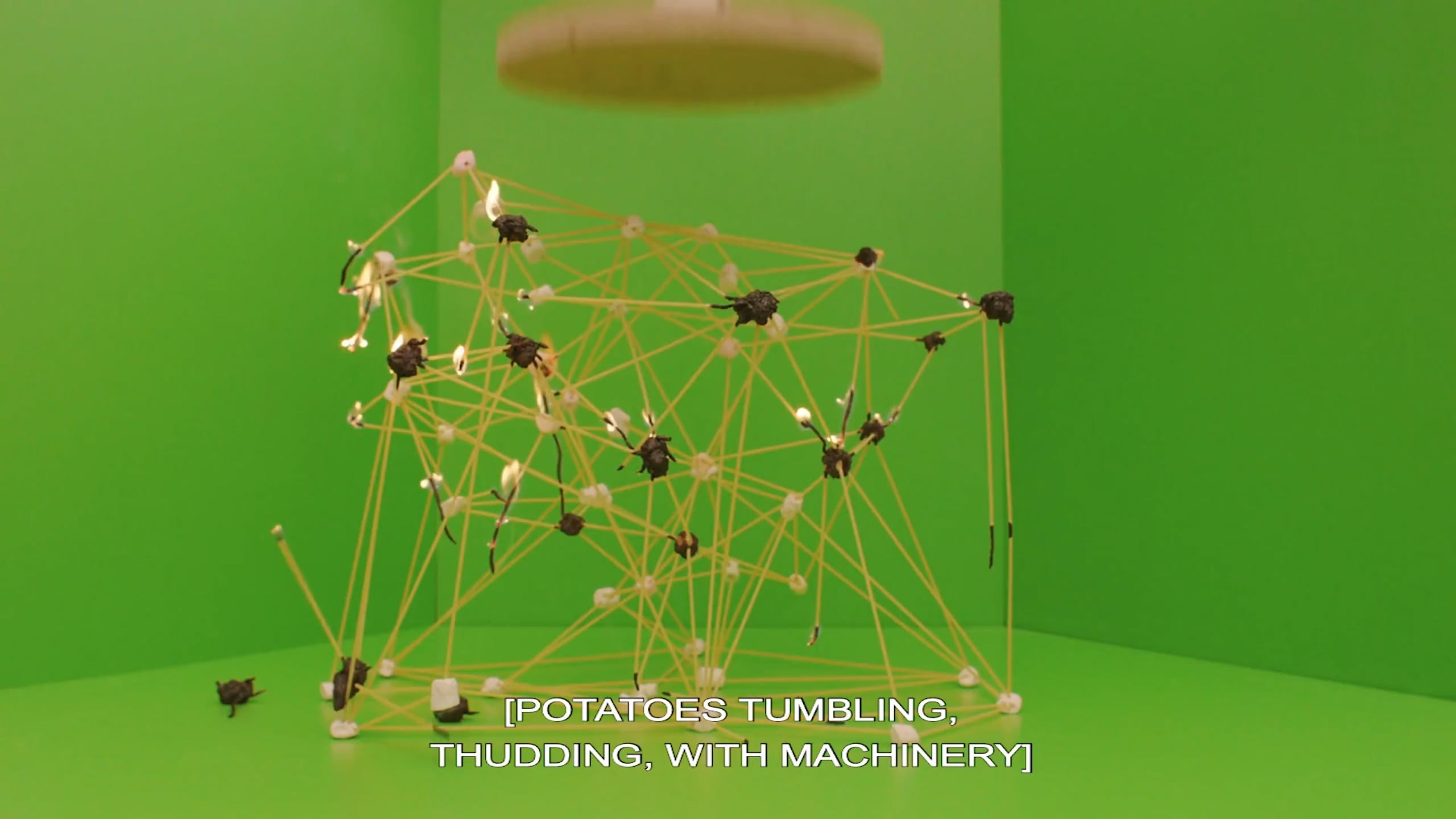 An elaborate, geometric structure in a green room is connected by marshmallows, some of which are on fire. Text on the image says [POTATOES TUMBLING, THUDDING, WITH MACHINERY].