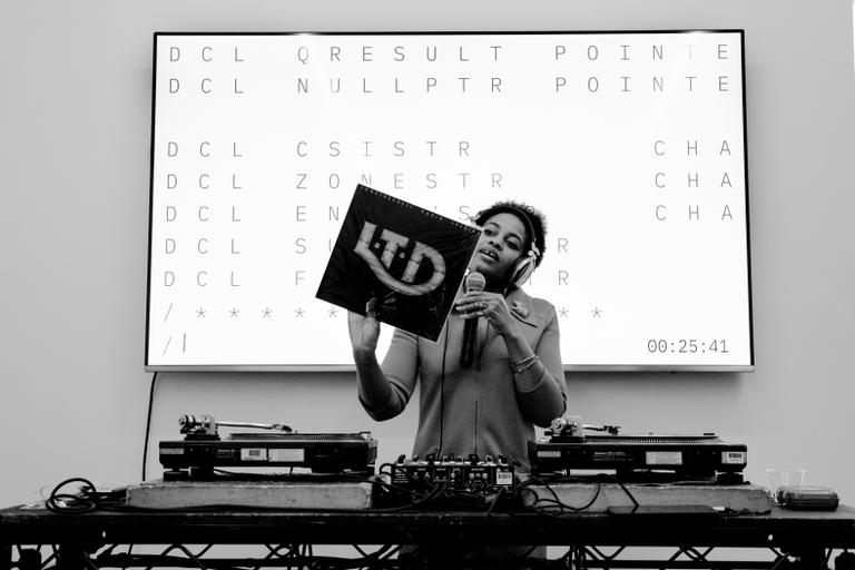 A person wearing headphones stands at a table with two turntables in front of a large video screen while holding a microphone and an album cover that reads LTD.