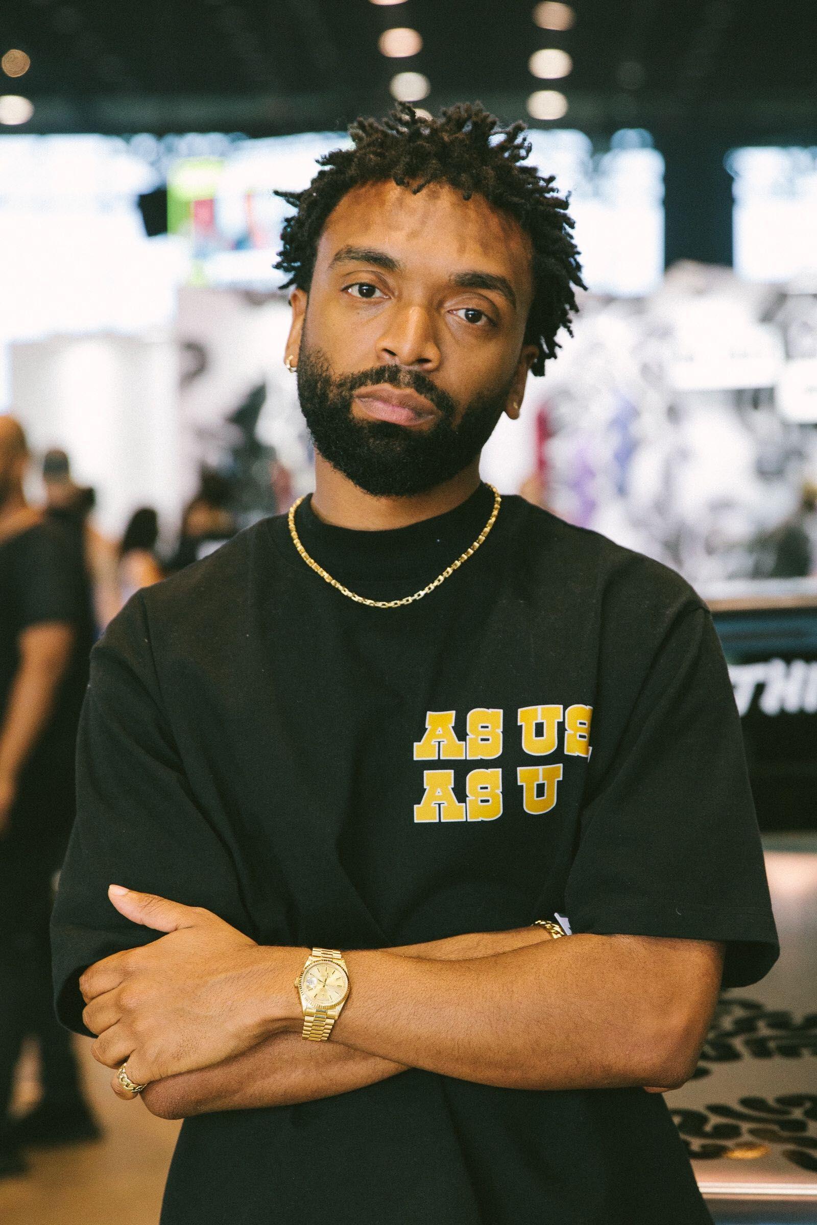 A person with short dreadlocks and a beard stands with arms crossed and head slightly tilted to their left while regarding the viewer with a serious expression.