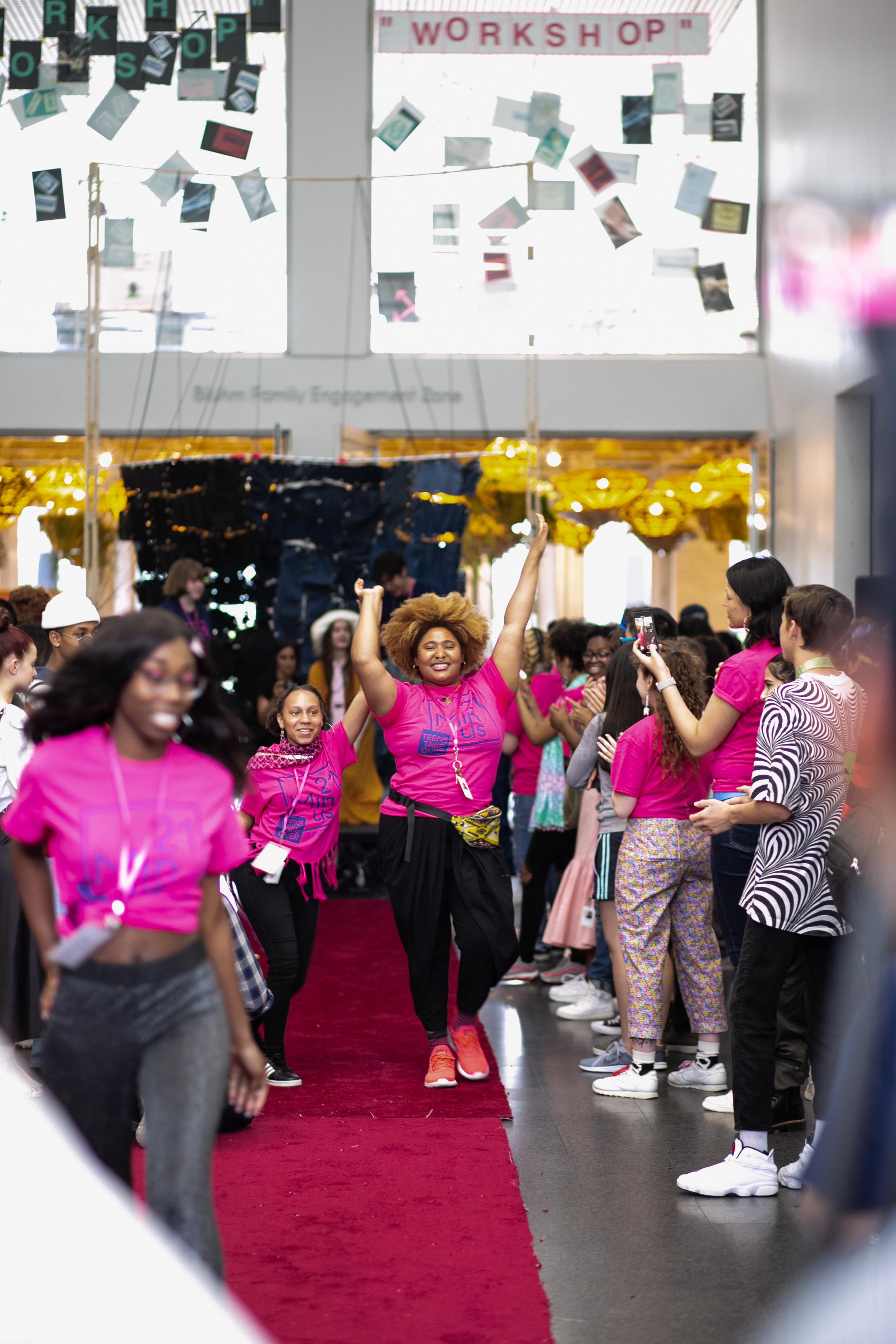 Young people in bright pink t-shirts walk jubilantly down a red carpet as onlookers watch and applaud.