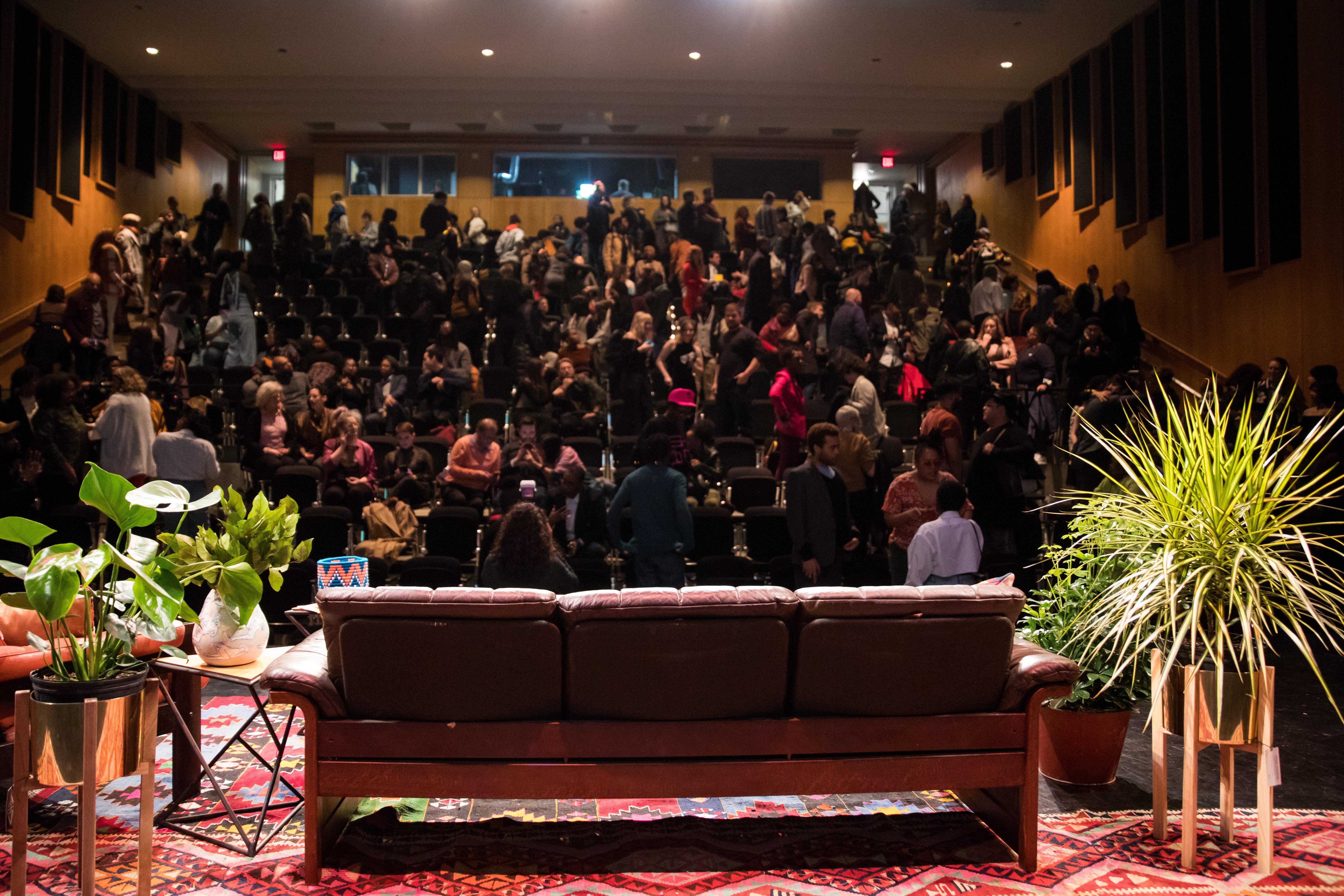 A view from the back of a stage set out towards a packed theater. The set contains a couch and several plants.