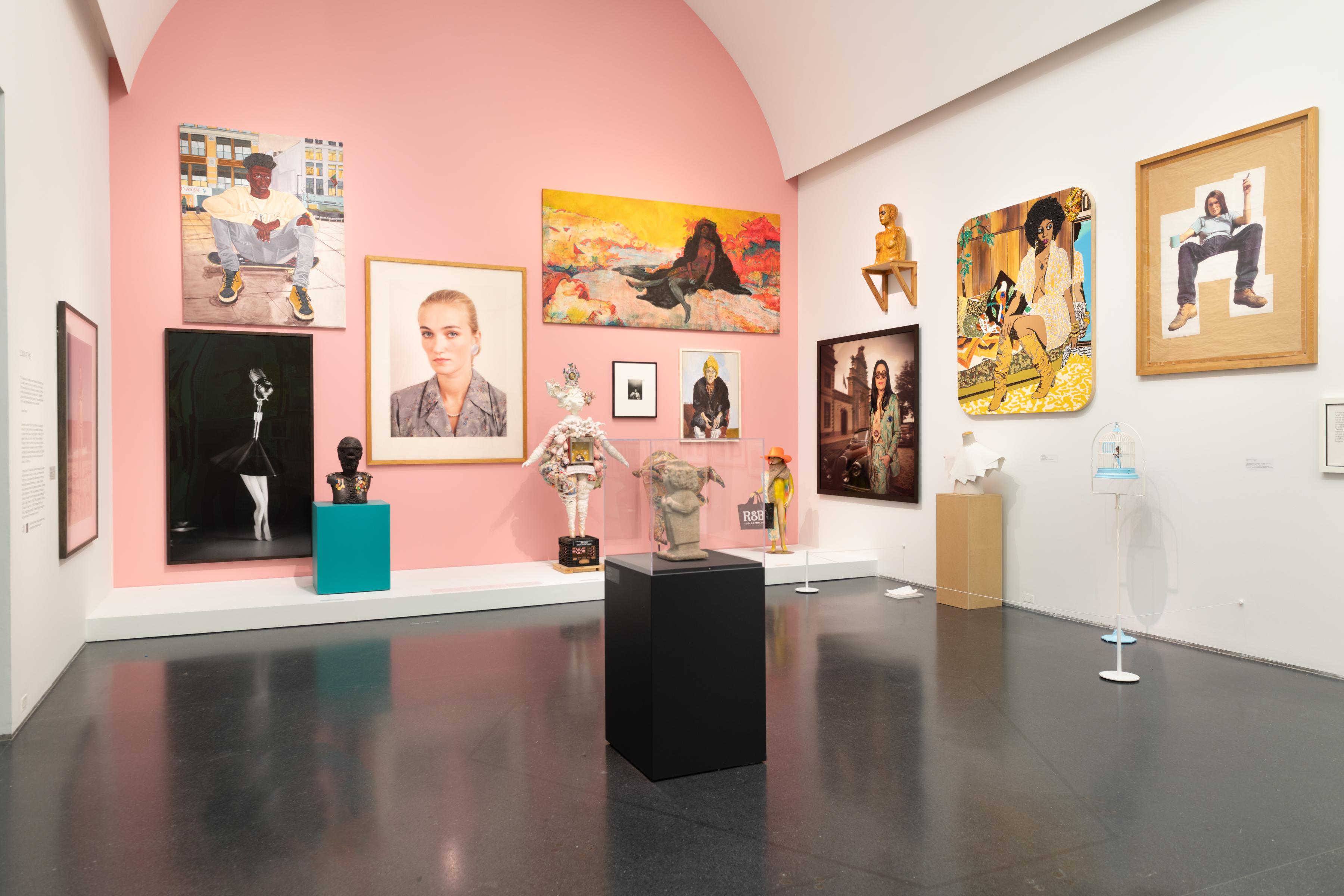 A pink, back wall is emphasized in a room with three walls shown. There are various paintings, photos and sculptures throughout the room.