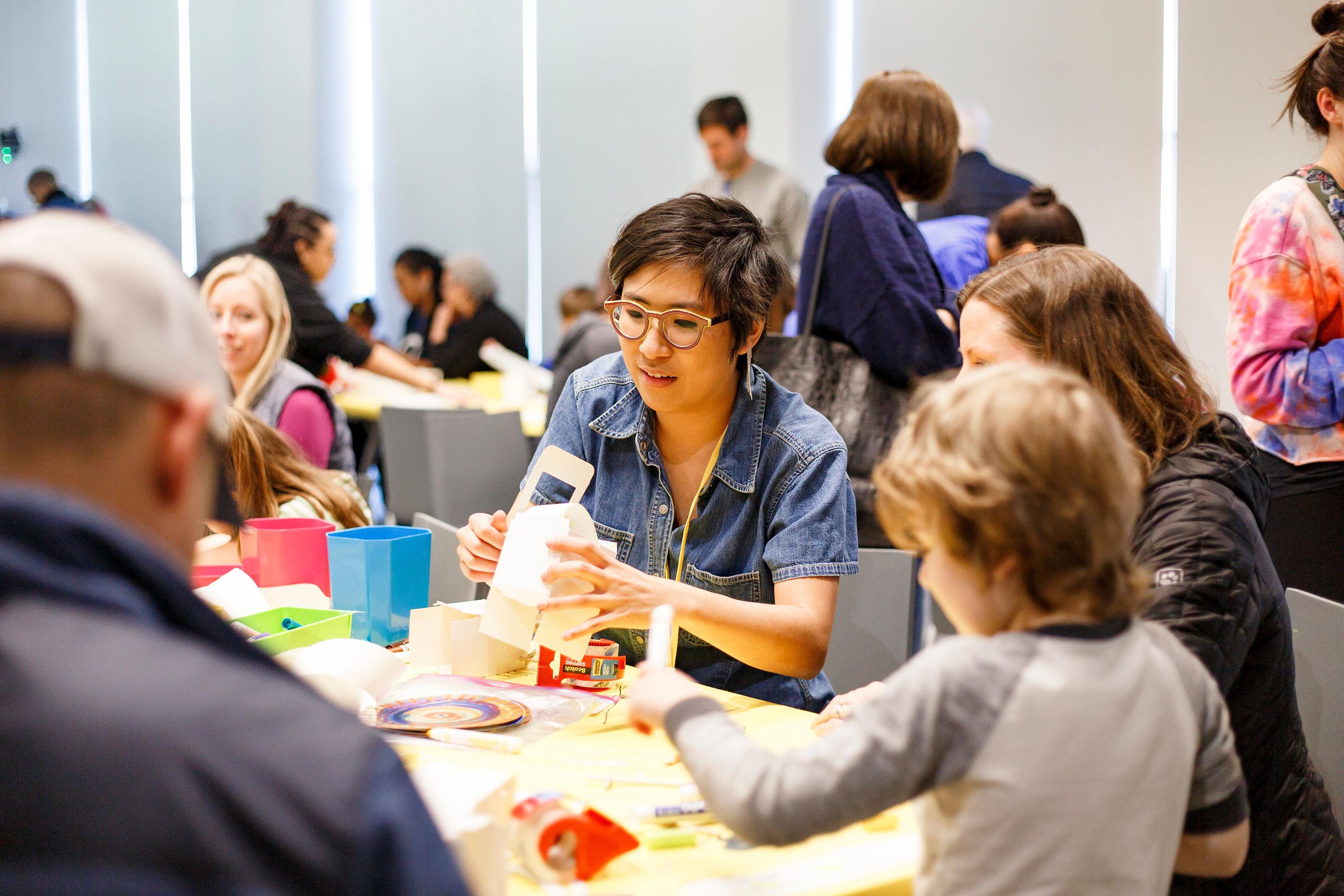 Smiling adults and children sit at tables creating objects with various material provided.