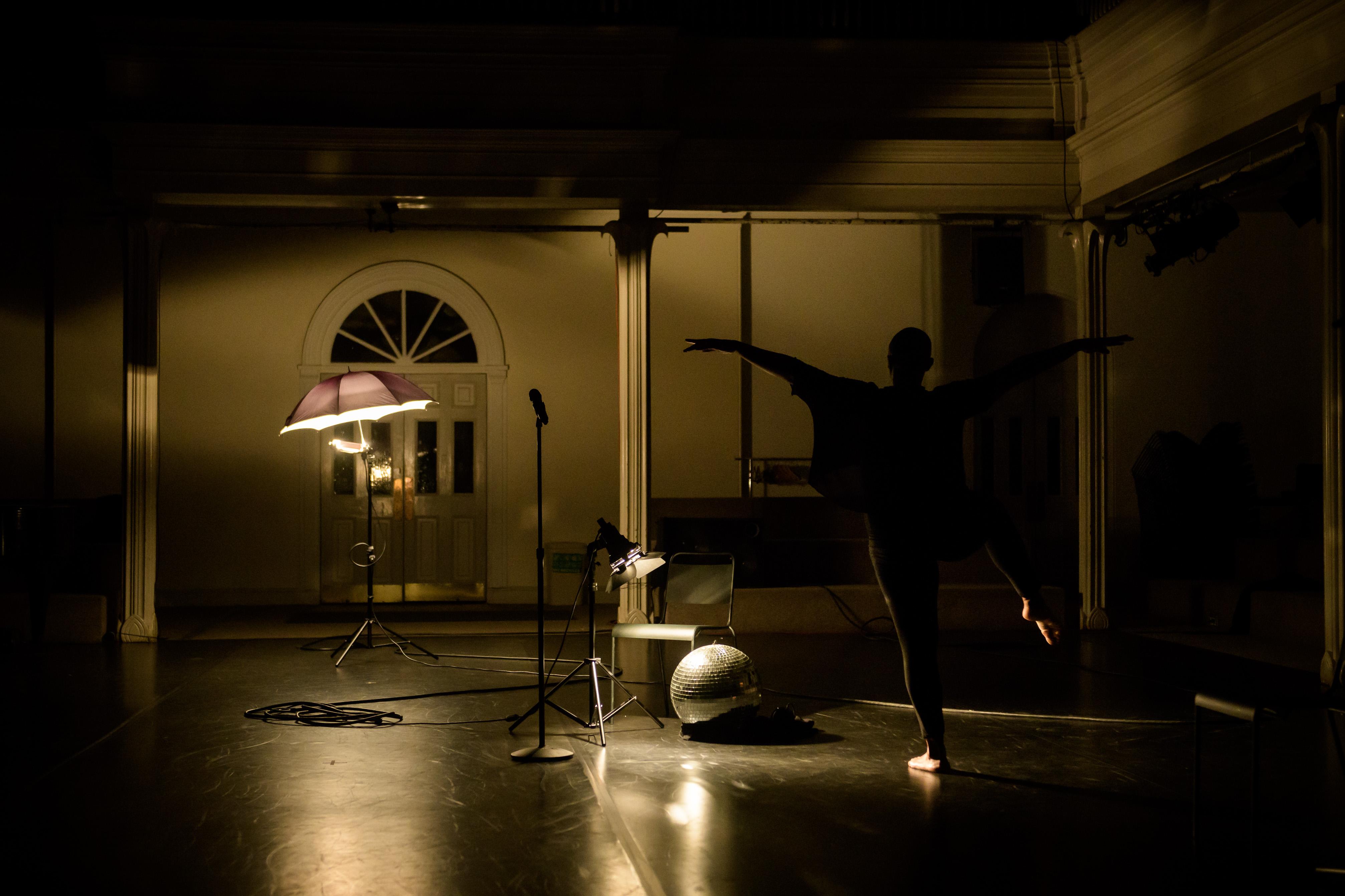 In a dramatically lit setting, a dancer in silhouette stands on one leg in arabesque with their arms extended like wings.