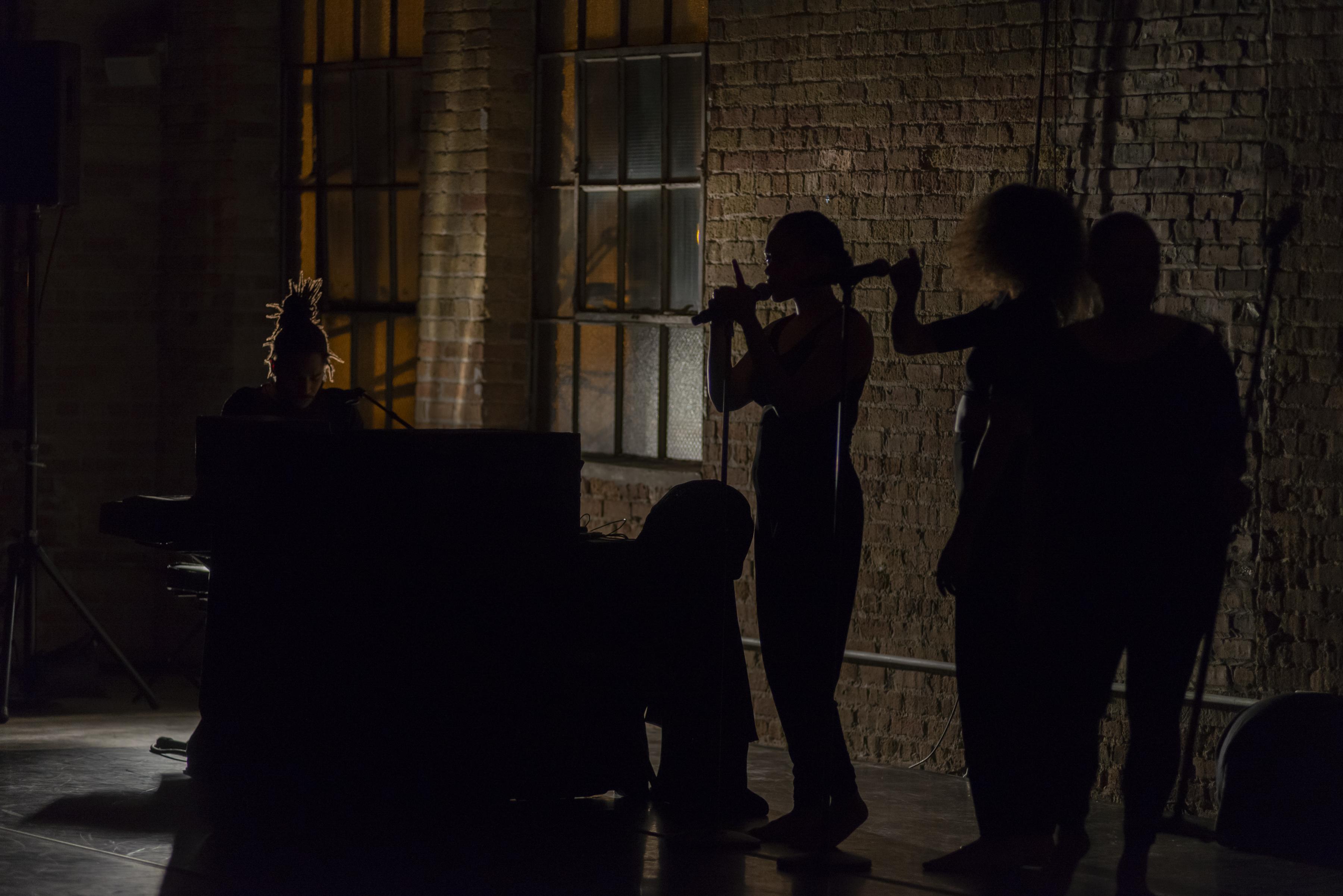 A group of performers are silhouetted against a brown brick wall.