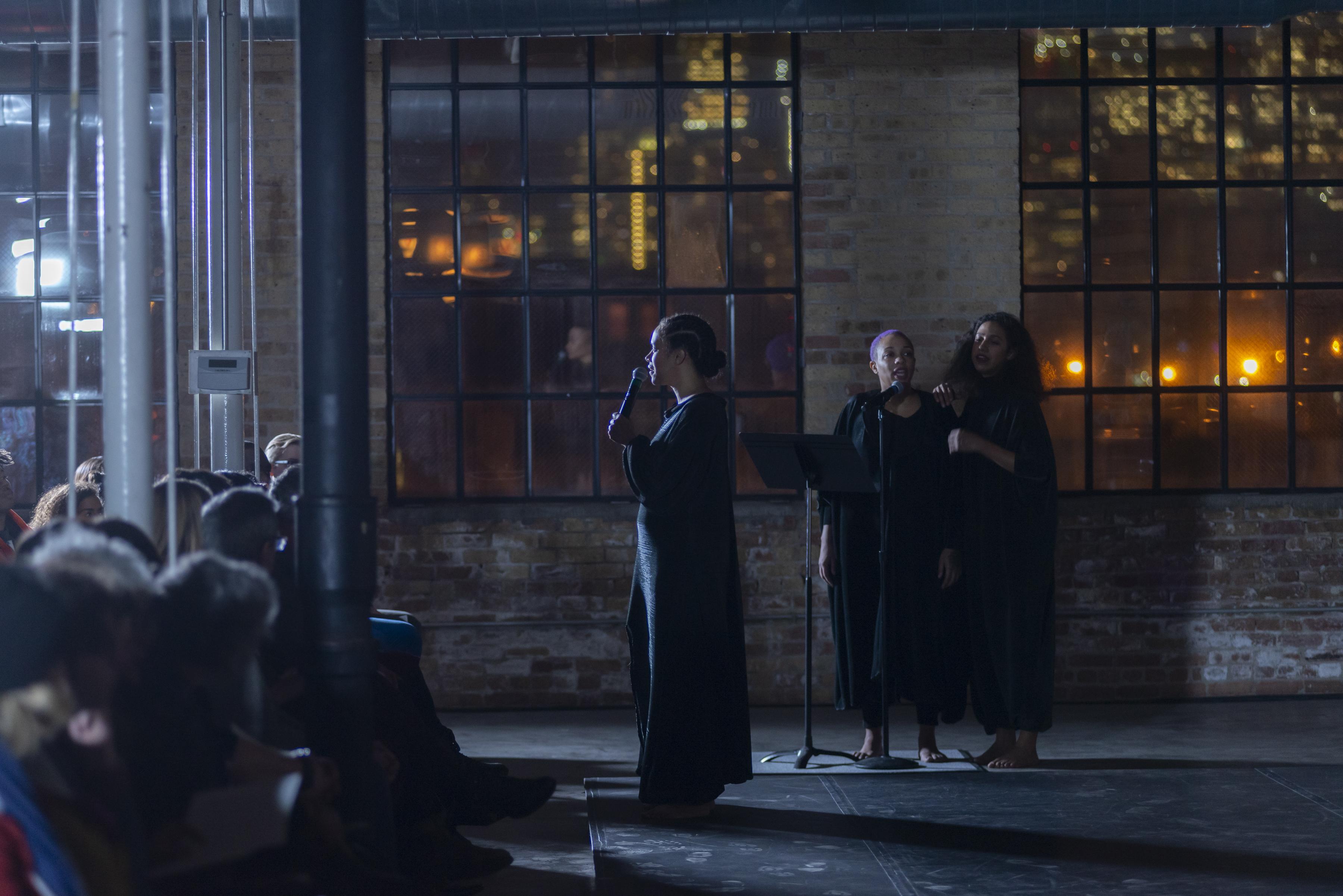 Before an audience, a person in a long, dark robe holds a microphone as two barefoot people in dark robes sing in the background.