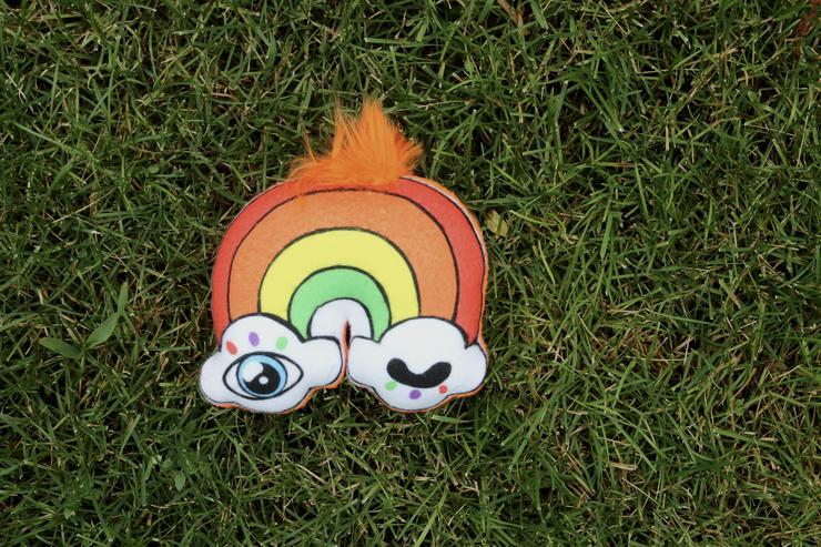 An orange rainbow with winky eye clouds on each end and a tuft of orange fluff on top lays on a bed of grass.