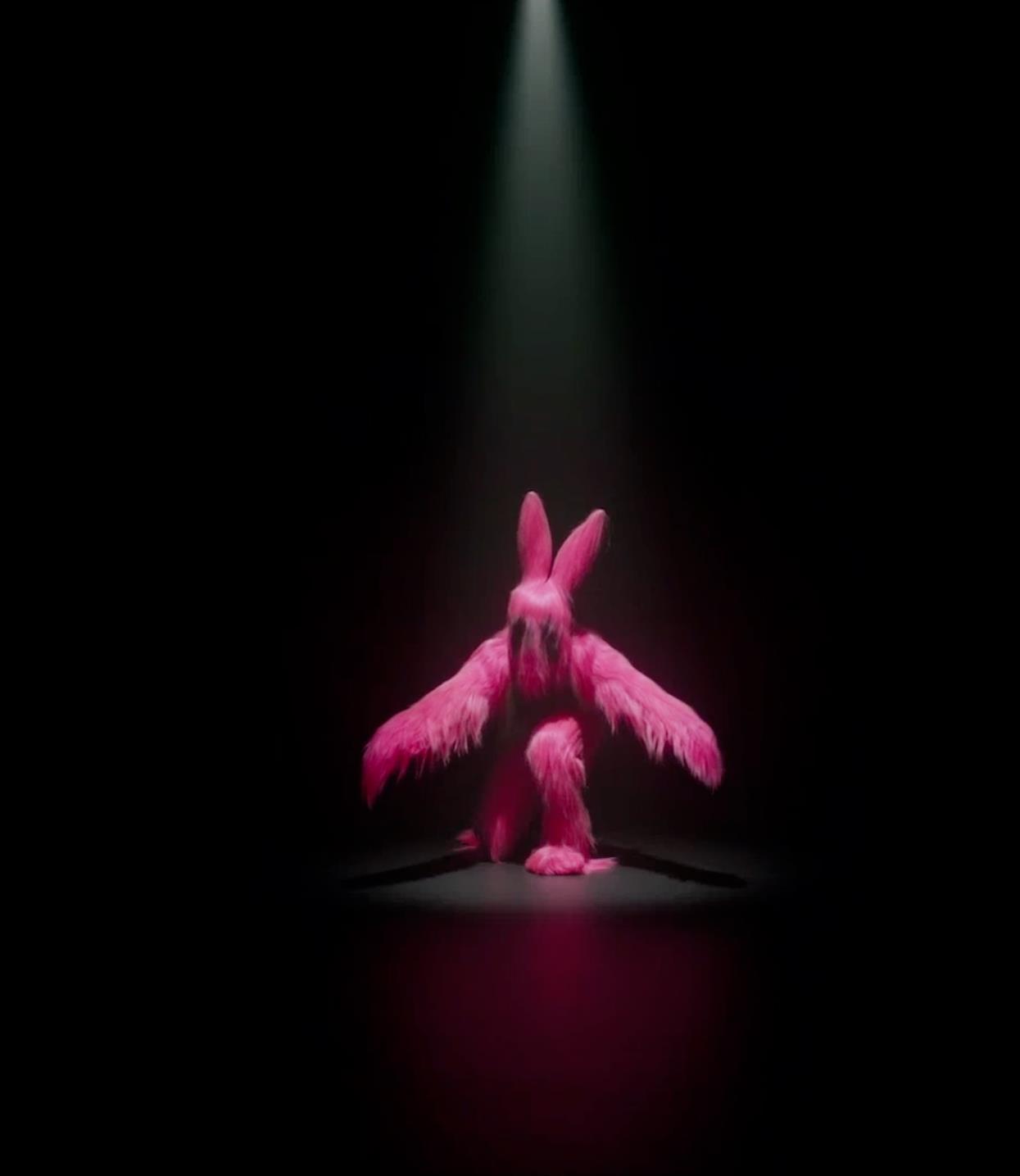 A performer costumed in a pink feathery bunny suit crouches with one leg forward, arms extended