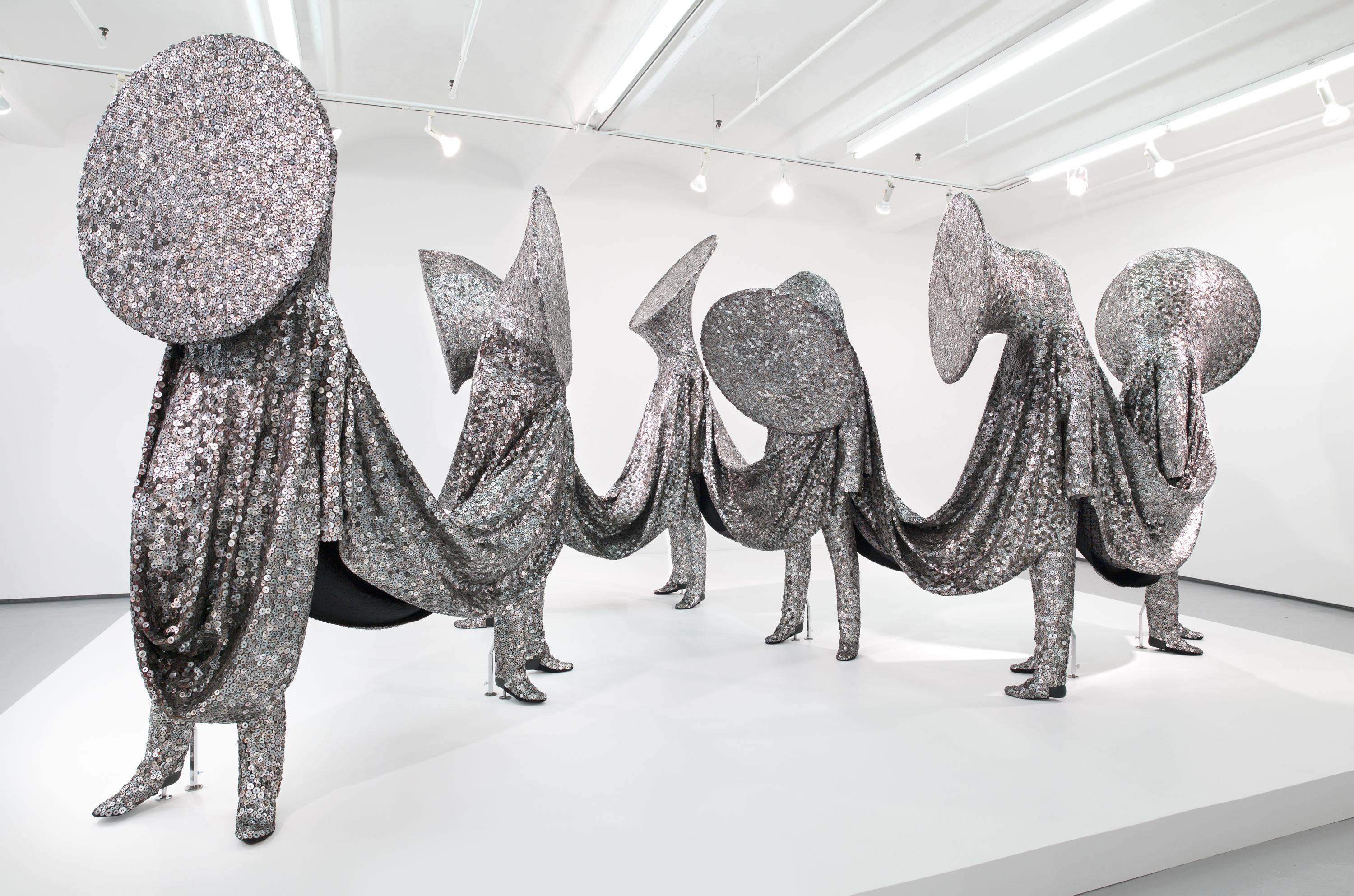a group of seven anthropomorphic sculptures with heads resembling flared horns are connected via draped fabric
