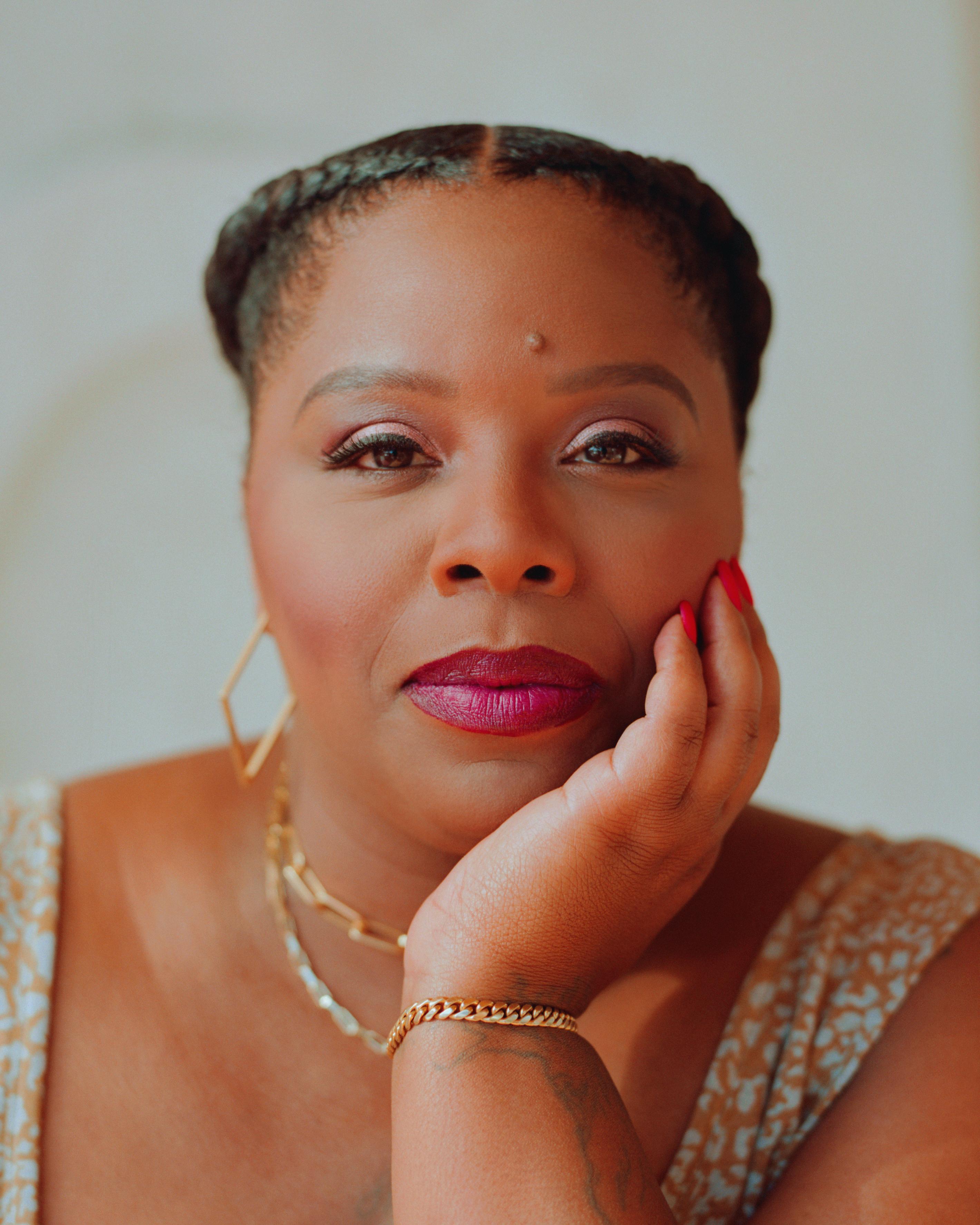 A Black woman with brown skin and brown hair pulled back in braids, her chin and cheek rests on her hand and she is wearing magenta lip color.