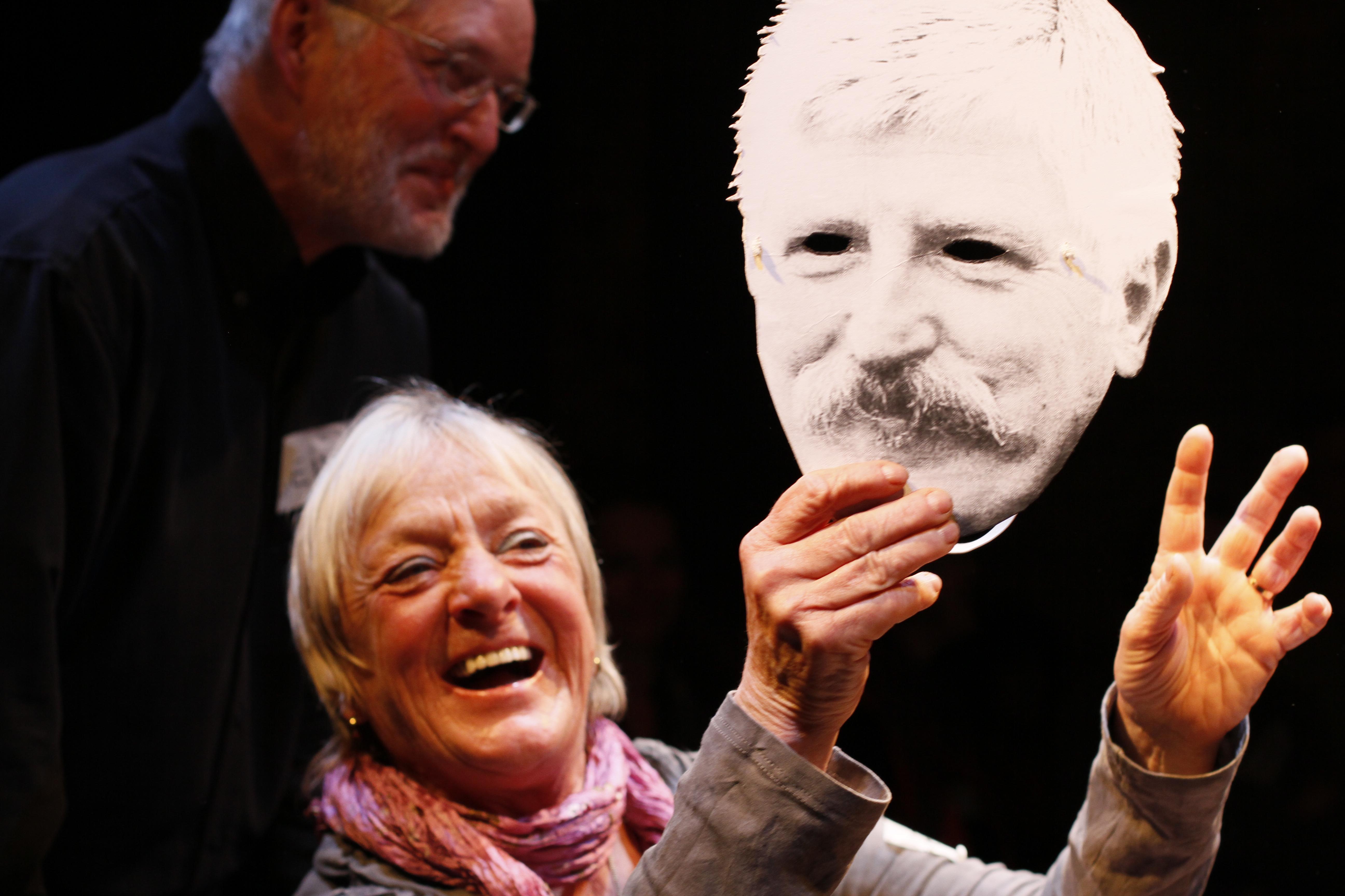 A woman holds a cardboard mask printed with the face of a mustached man