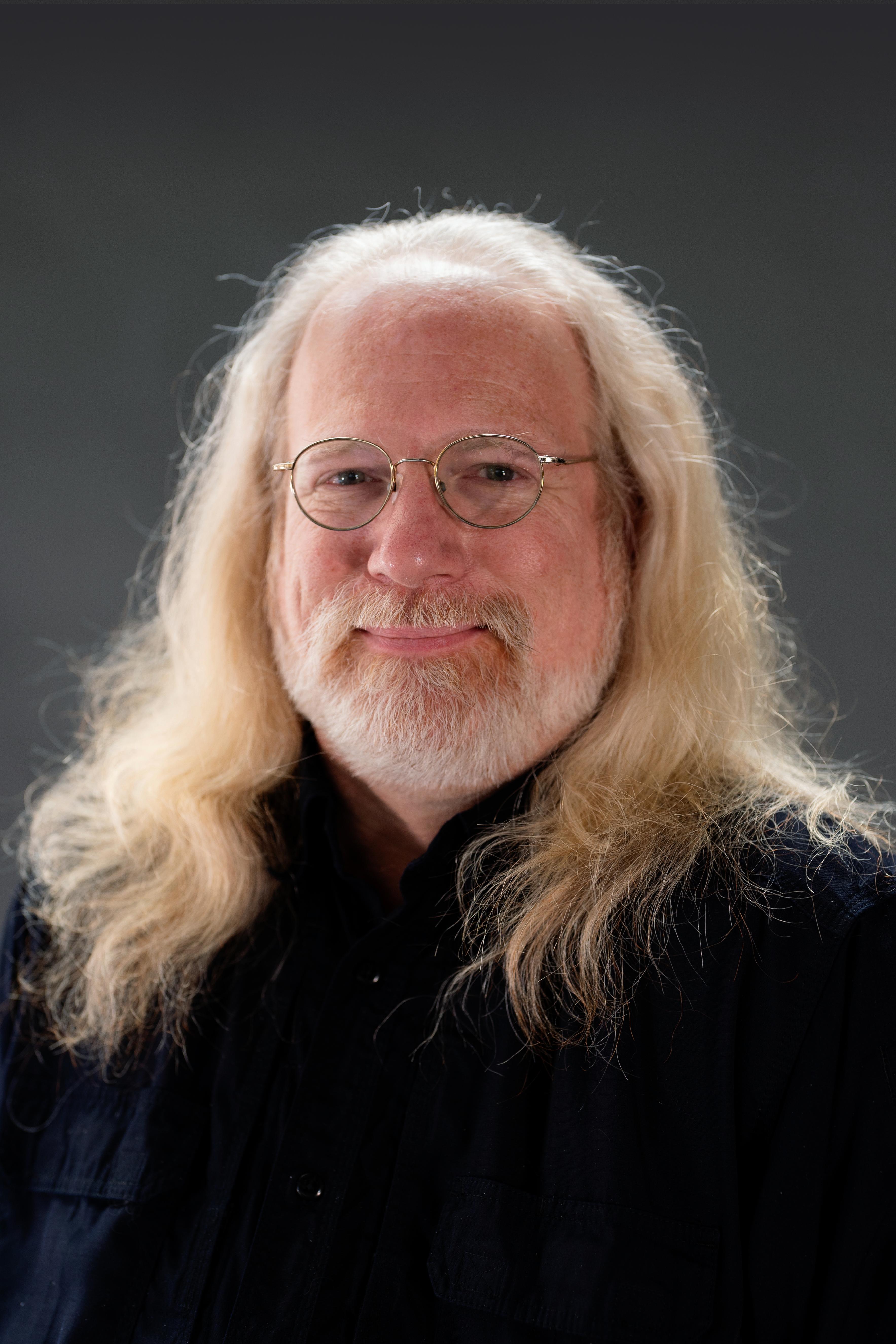 Michael Maag headshot. He is a white man with flowing white-blonde hair and a short beard. He wears wire-rimmed glasses and a dark blue collared shirt as he smiles warmly.