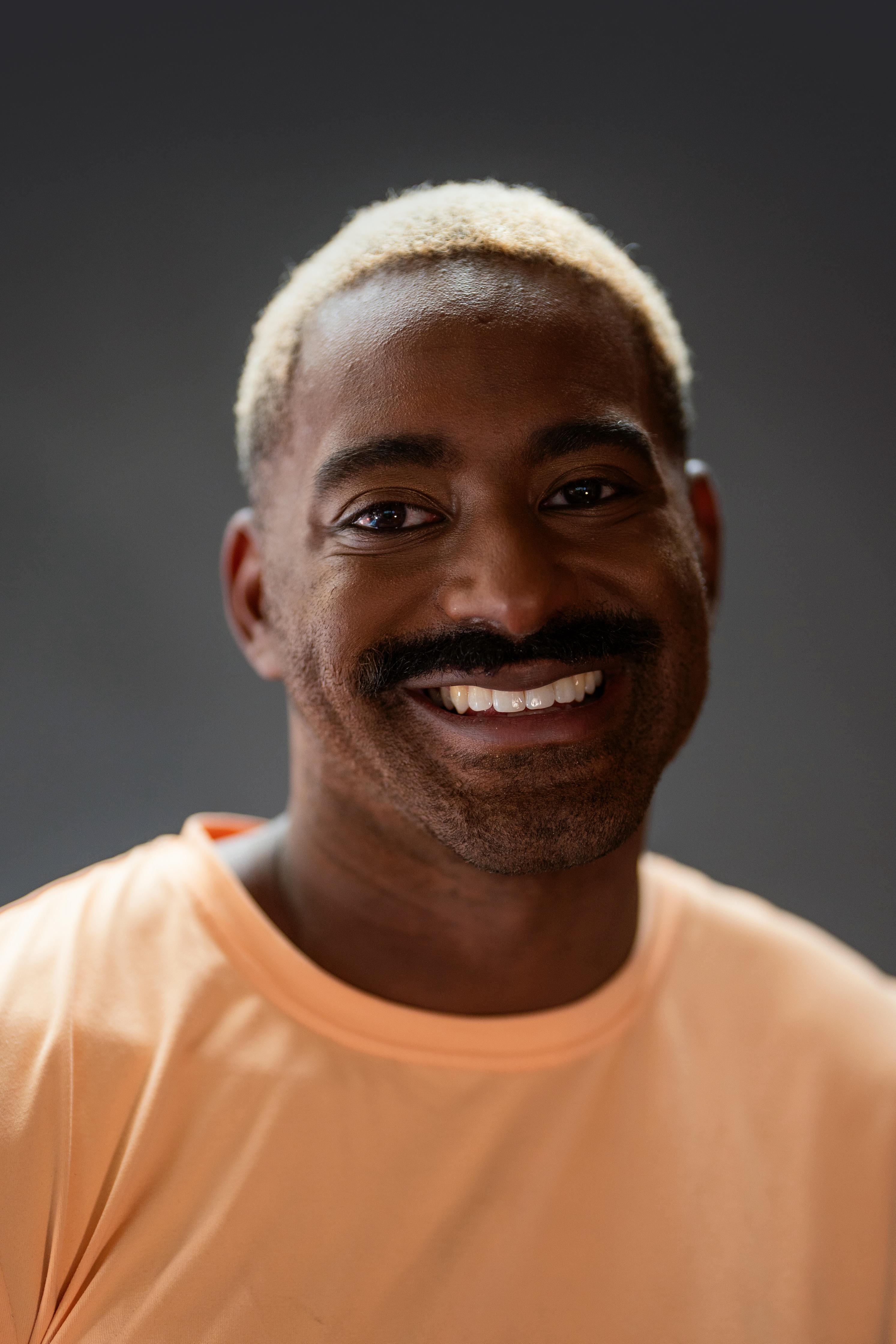 Jerron Herman headshot. He is a dark-skinned Black man with a big smile, dark moustache, and bright blonde hair; he wears a soft yellow T-shirt. Photo: Robbie Sweeny.