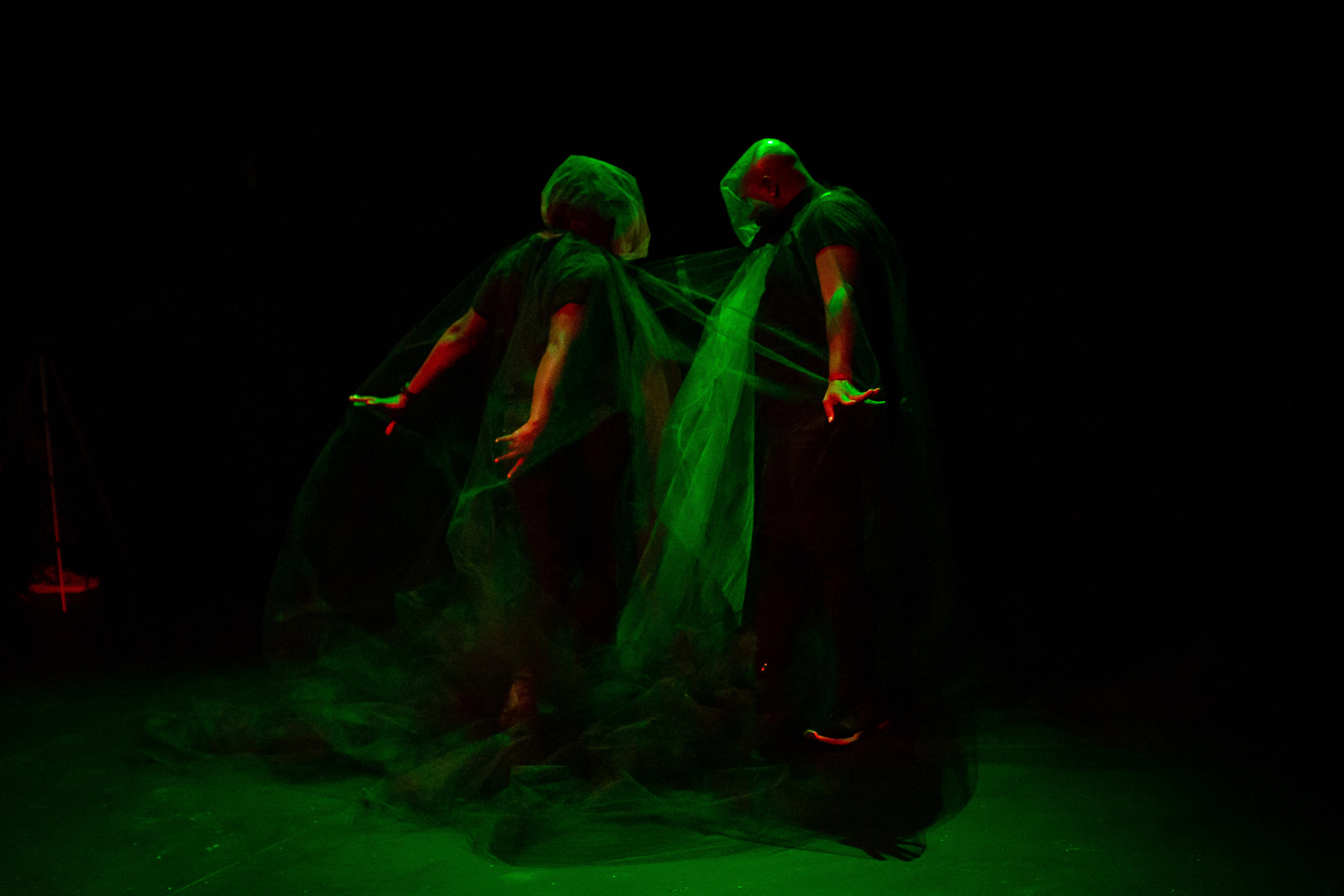Two shrouded figures stand on stage under soft green light