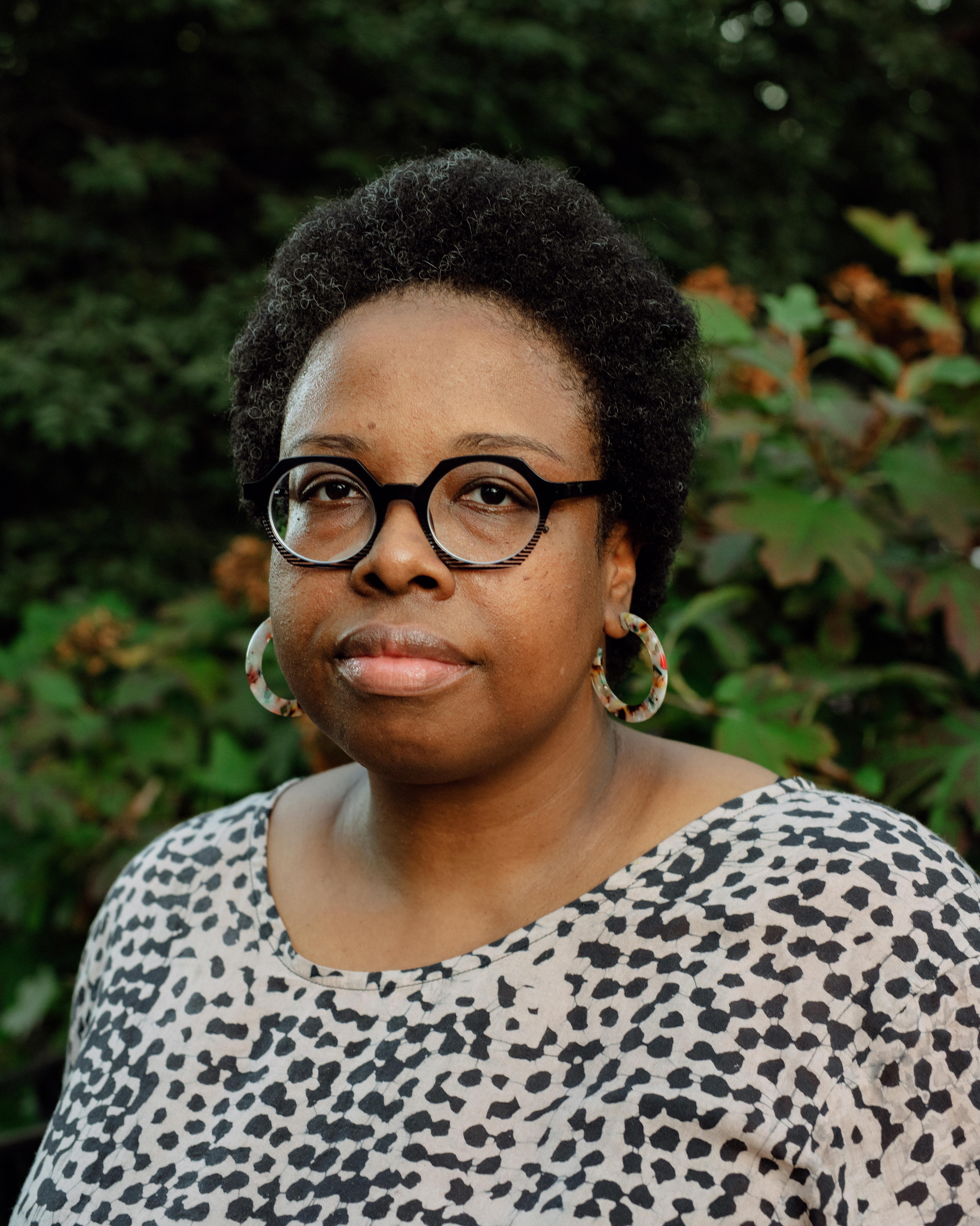 A headshot of a woman wearing glasses and a patterned shirt in front of a forest backdrop