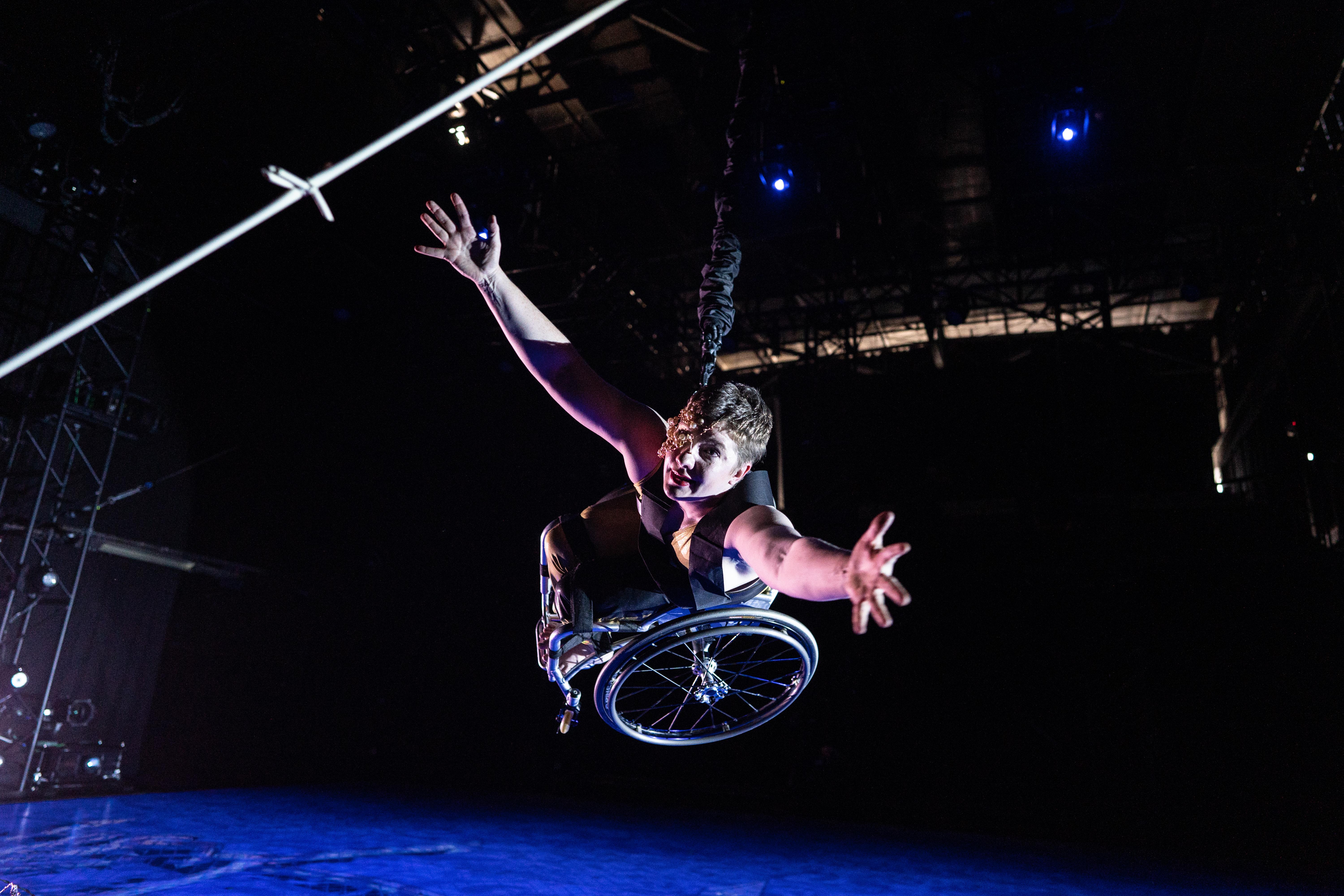 Laurel Lawson, a white woman with very short cropped hair, bounds toward the camera: wheelchair high off the ground, arms open and reaching. A mask of delicate gold wire and lustrous pearls covers half her face. The stage floor below her is drenched in blue light; the same light gives her pale skin a light violet glow. A strand of silver barbed wire appears, close up, in the upper corner.