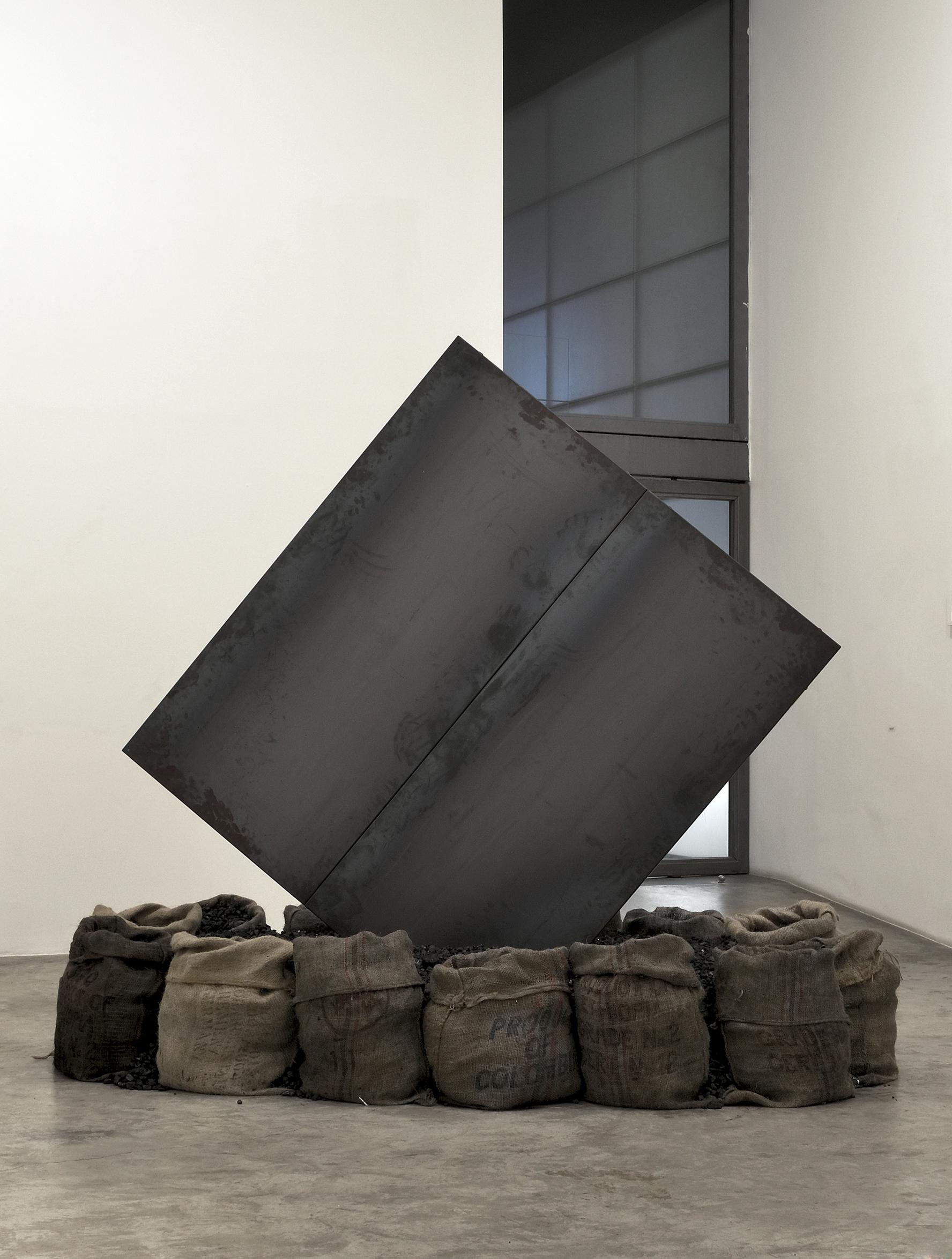 sculpture of square steel plate rotated and standing on point surrounded by burlap sacks filled with coal
