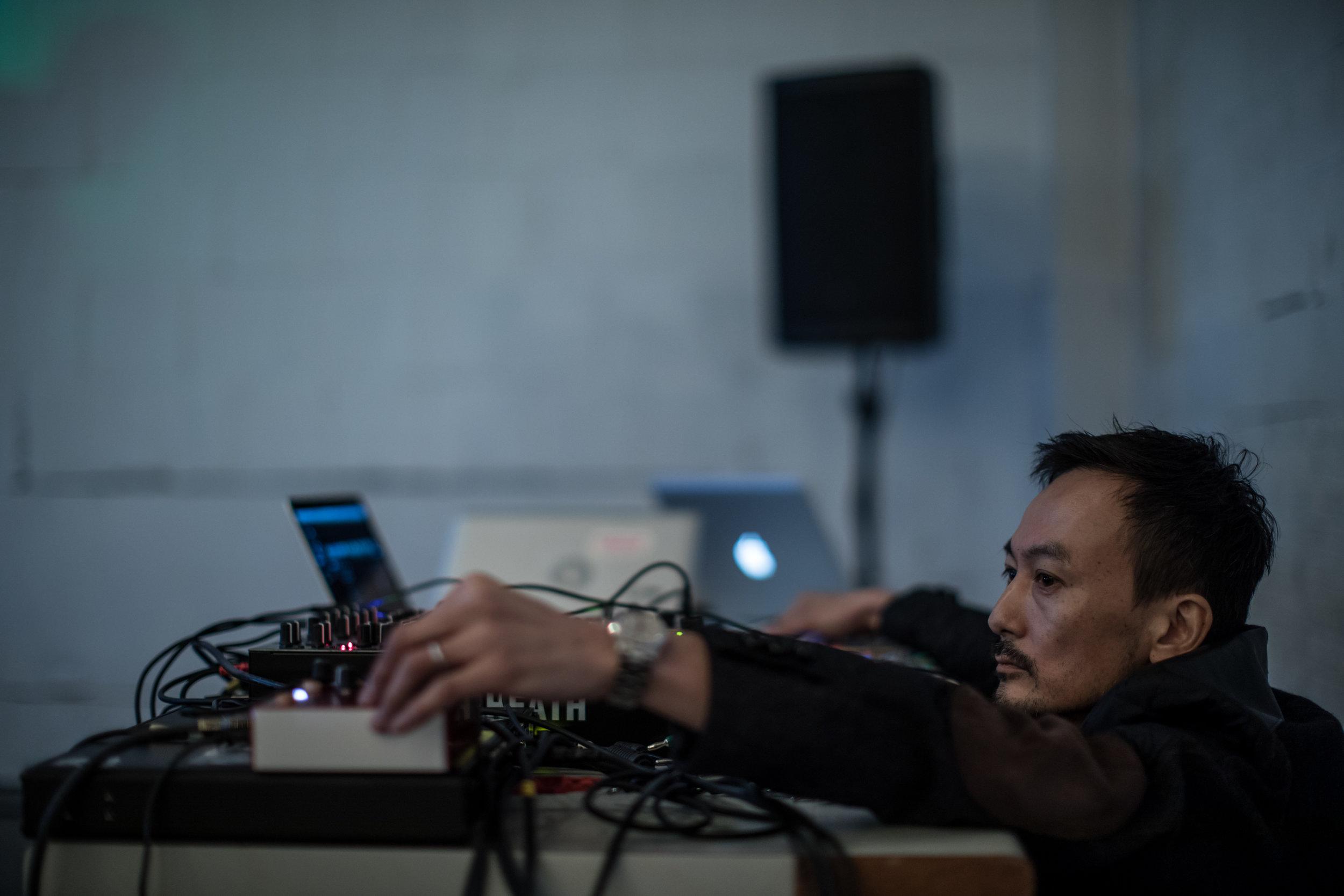 Kiku Hibino crouches at a table filled with audio equipment making adjustments