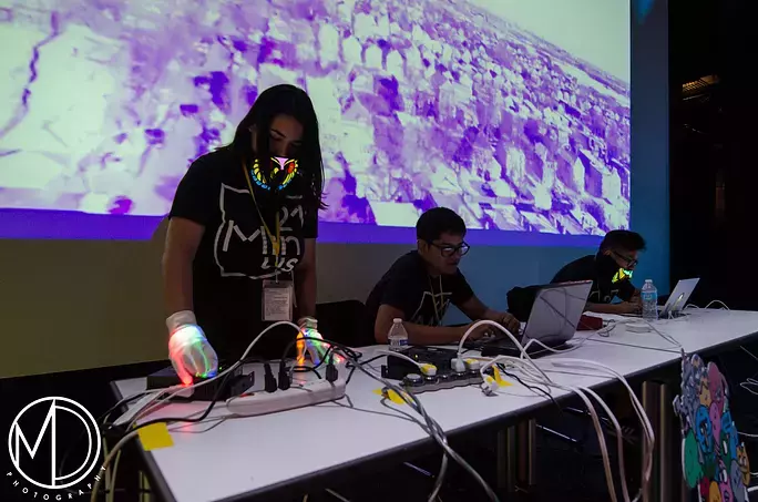 Three individuals adjust electronics and laptops in a darkened room in front of a projected image.