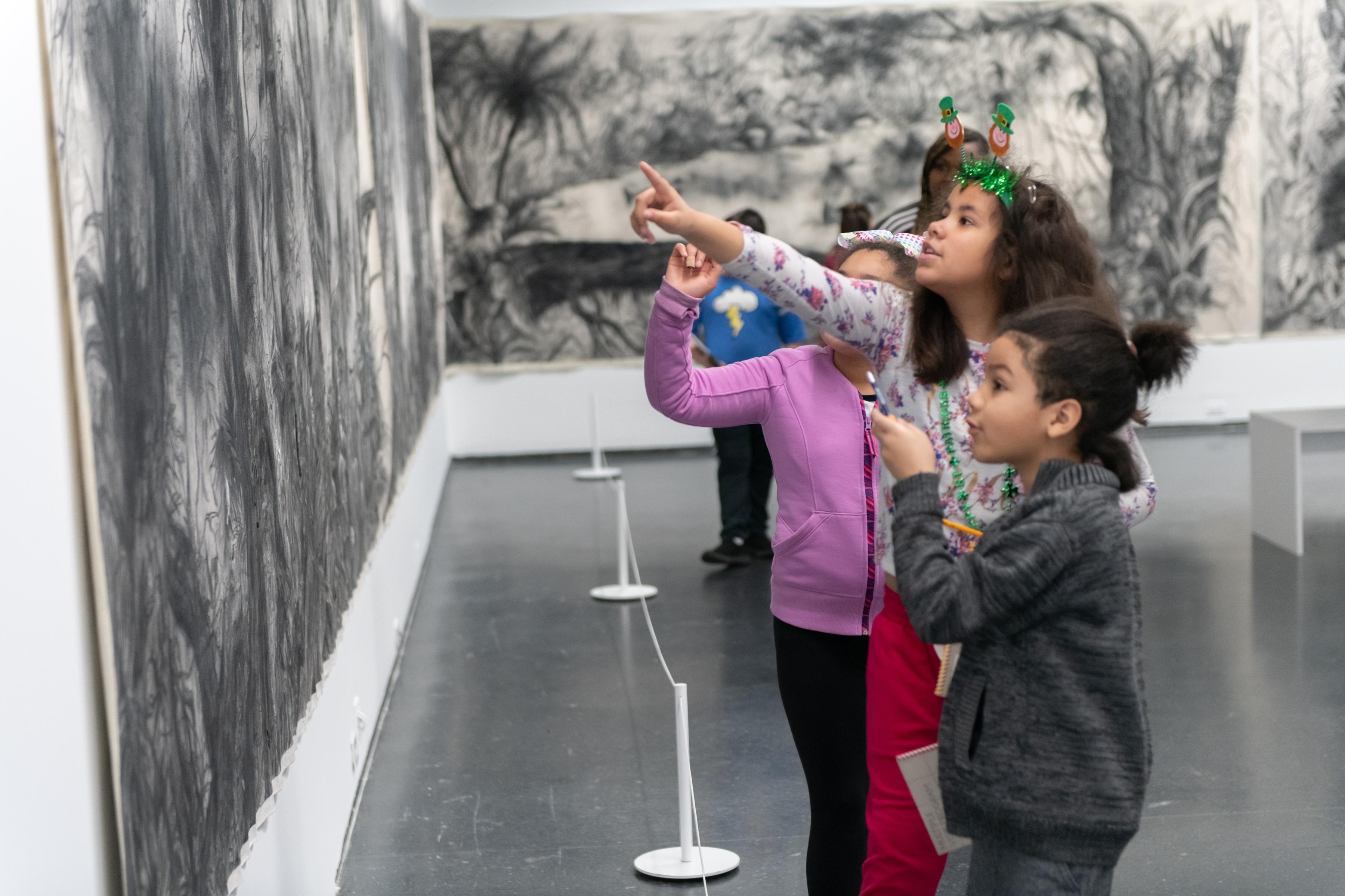 Los estudiantes miran y señalan un dibujo en blanco y negro de árboles tropicales y follaje que se muestra en dos paredes.