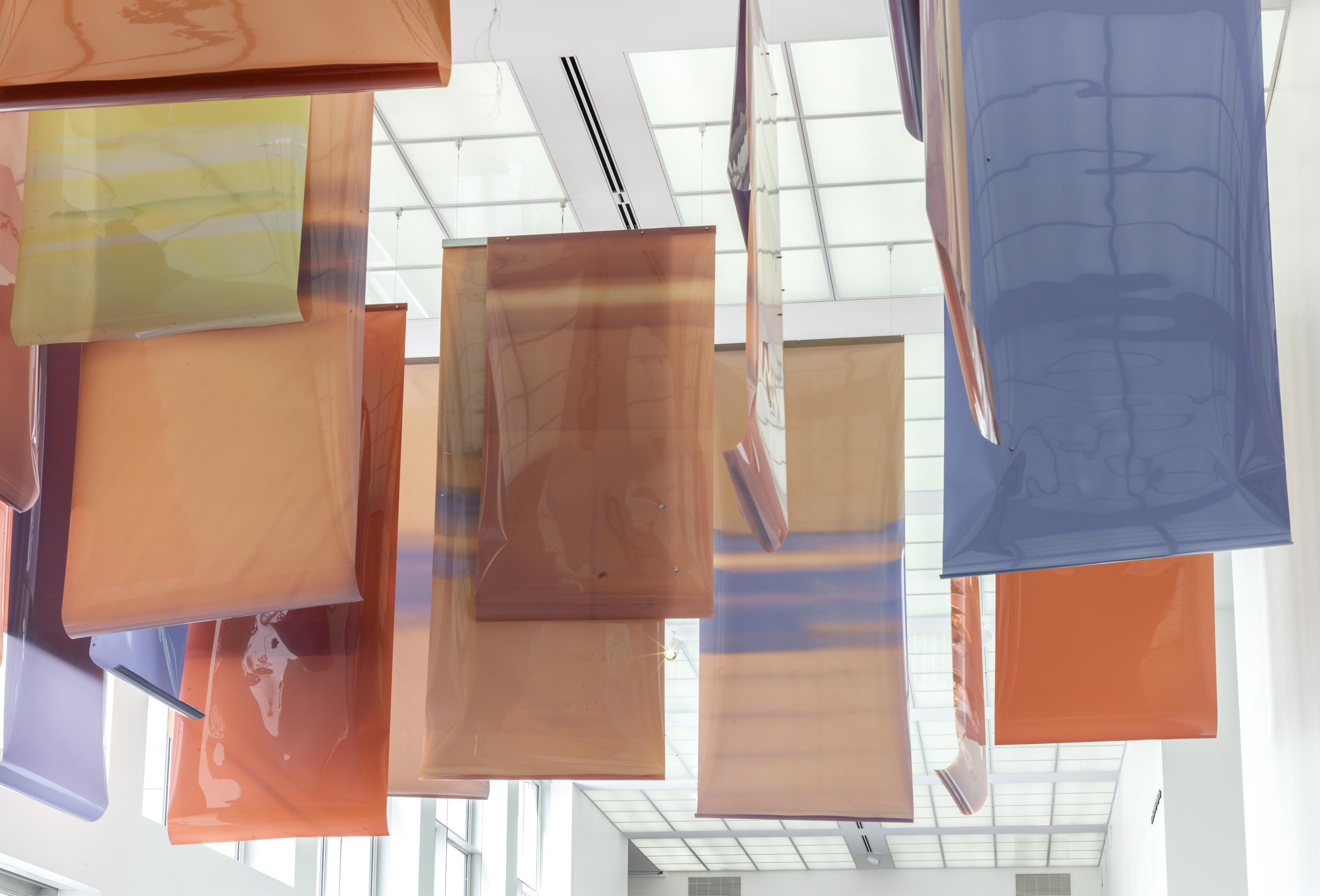 Large pieces of plastic in varied shades of blue, orange, peach, and yellow hang from the ceiling of an airy, white space.