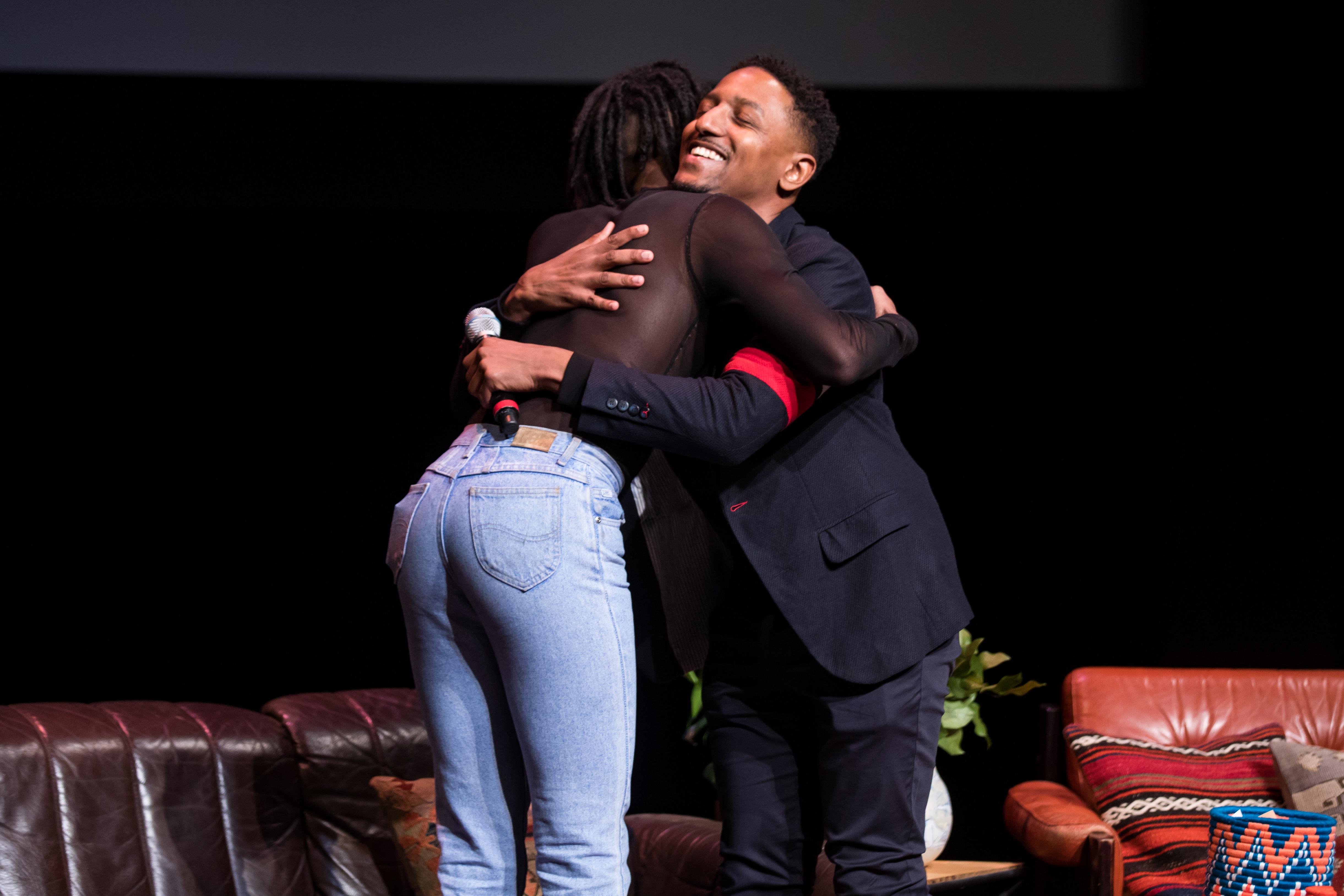 Two people embrace on a theater set. One holds a microphone.