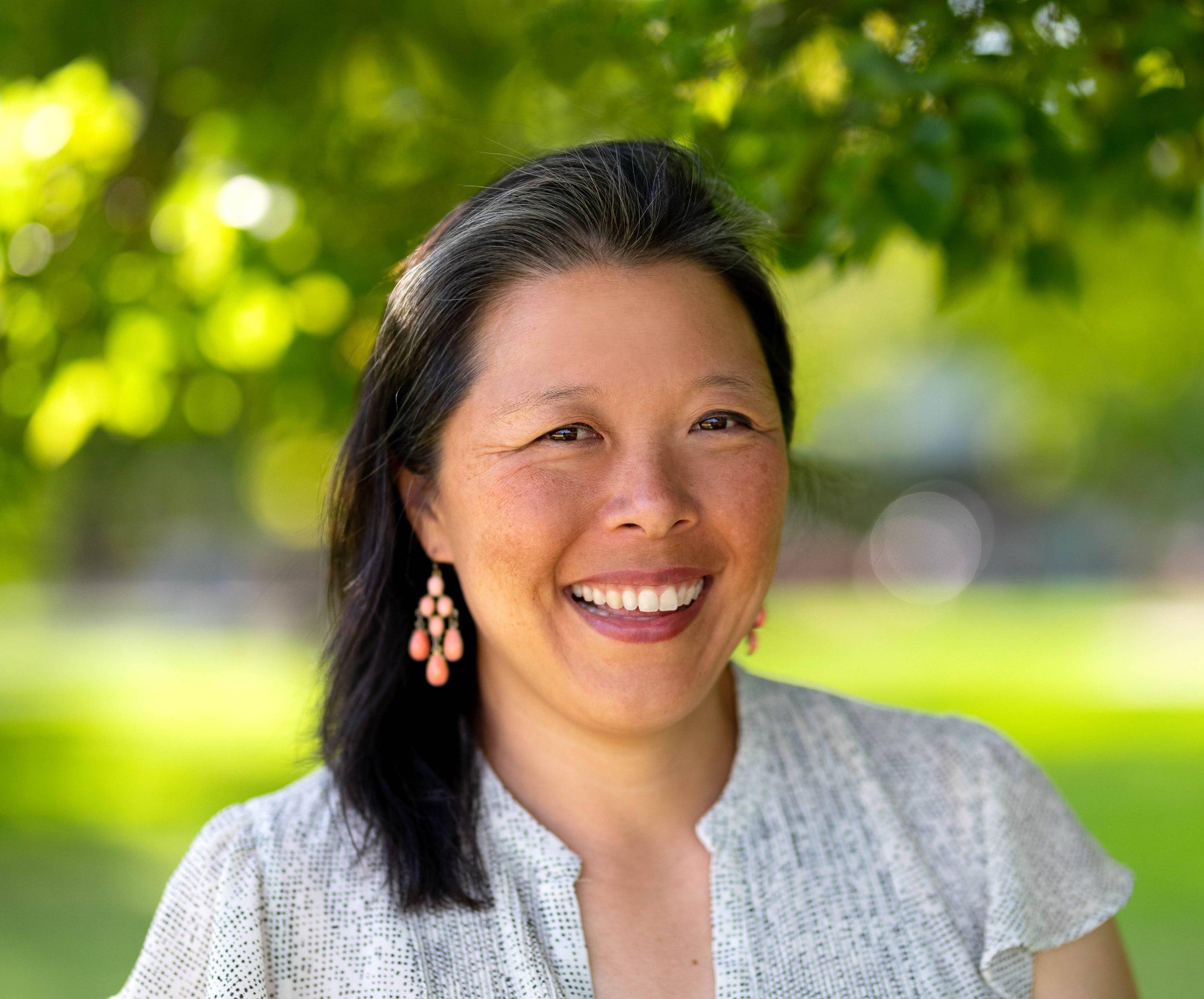 Portrait of a woman of Asian descent smiling at the camera. The background is blurred but appears to be outdoors.