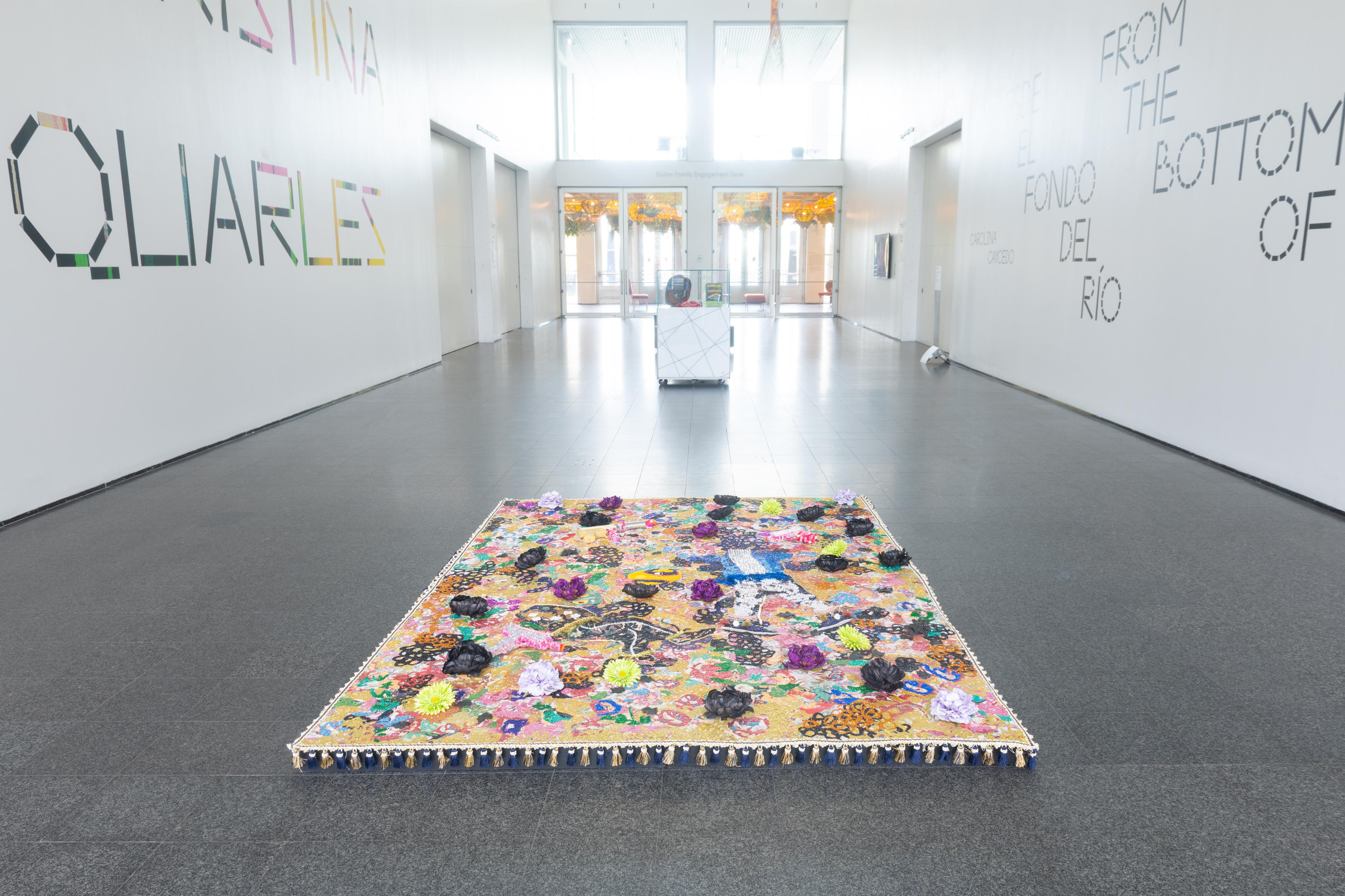 A colorful 3D rectangular rug sits in the middle of a large, white-walled room.