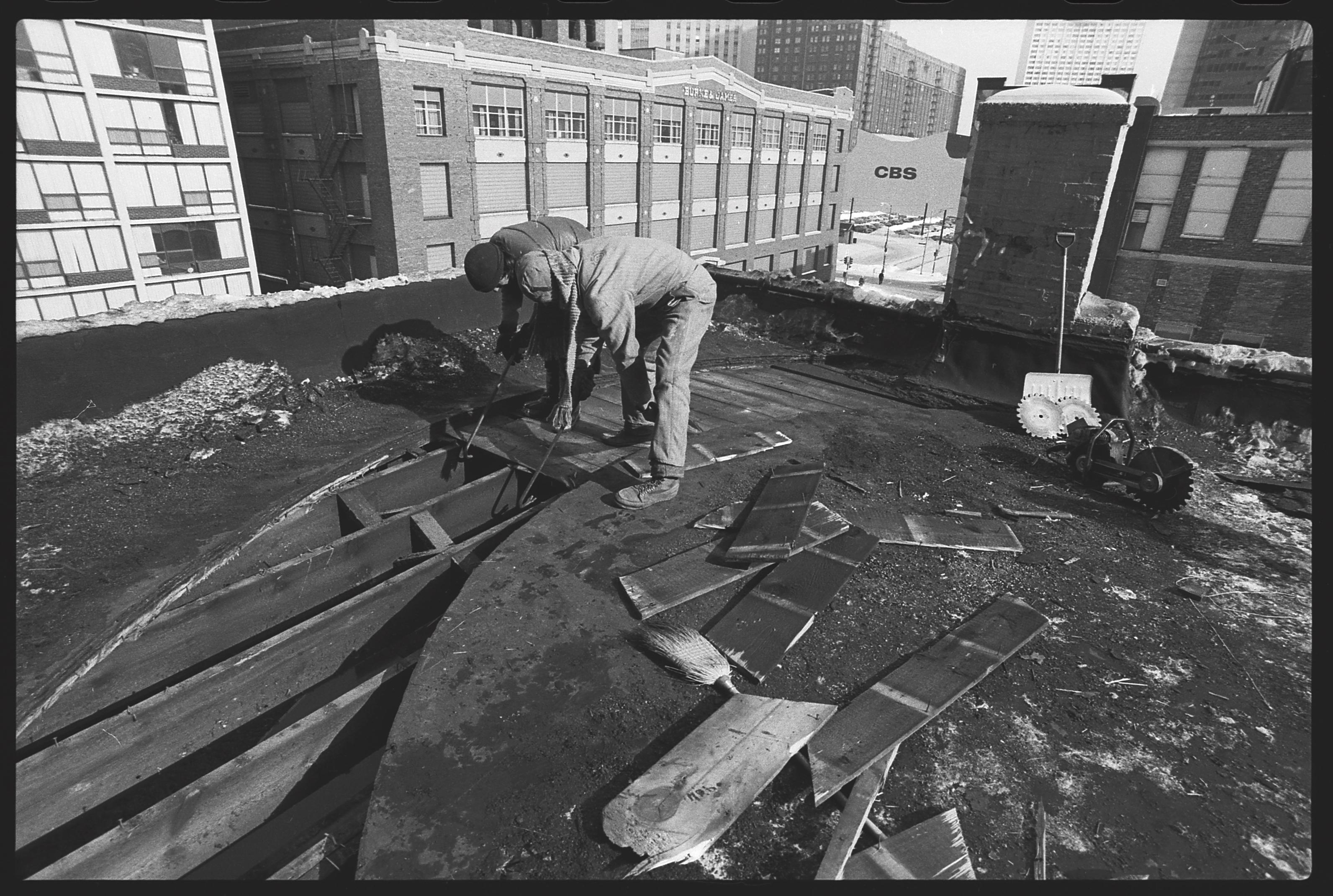Two people use crowbars to remove boards from the roof of a building in a curved manner. The rafters below are exposed in the part closest to the foreground.