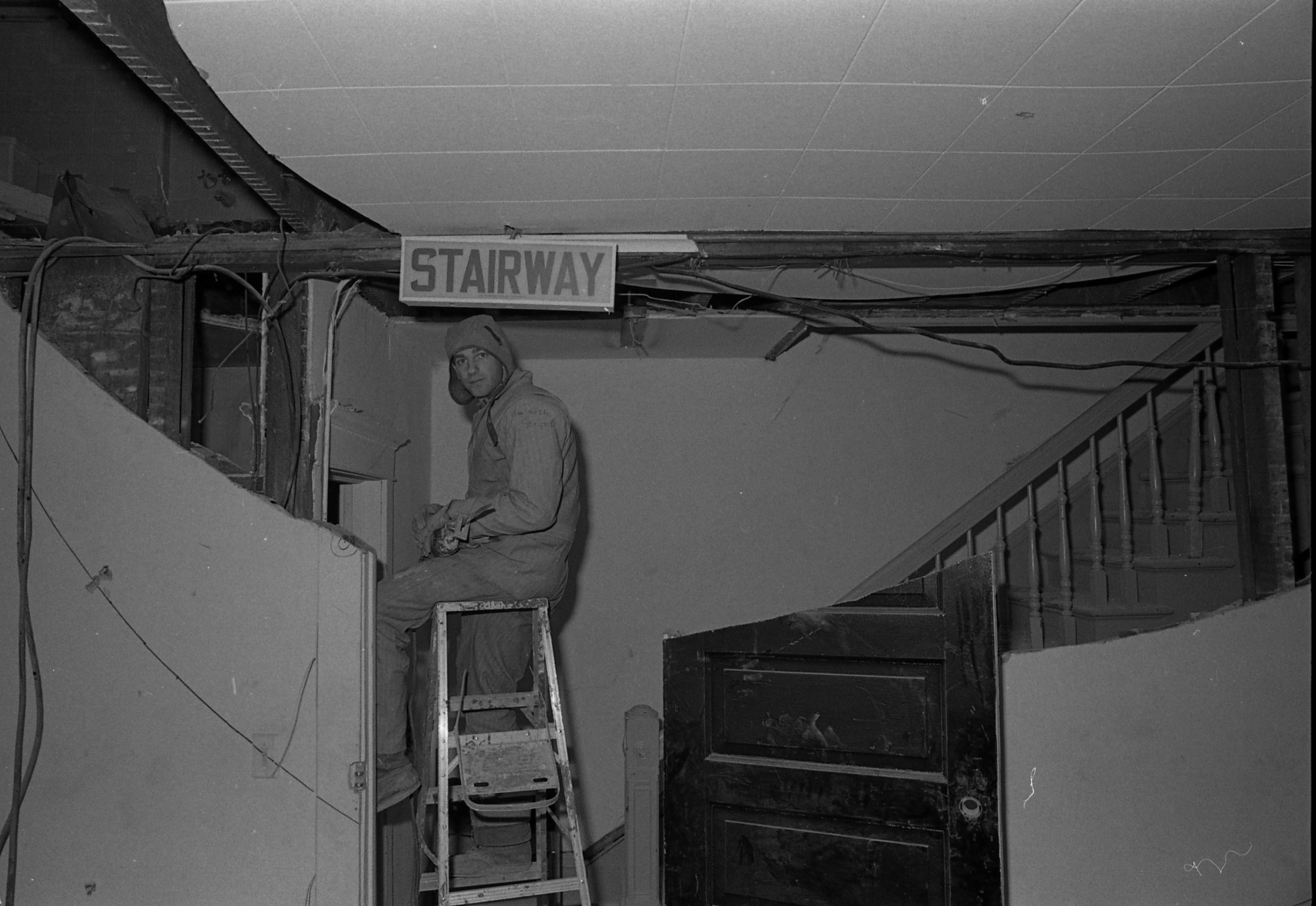 A man sits on a ladder underneath a sign that reads Stairway. The wall and door are cut in a way that they form a curve. His shirt has a handwritten phrase on it reading I'm with stupid.