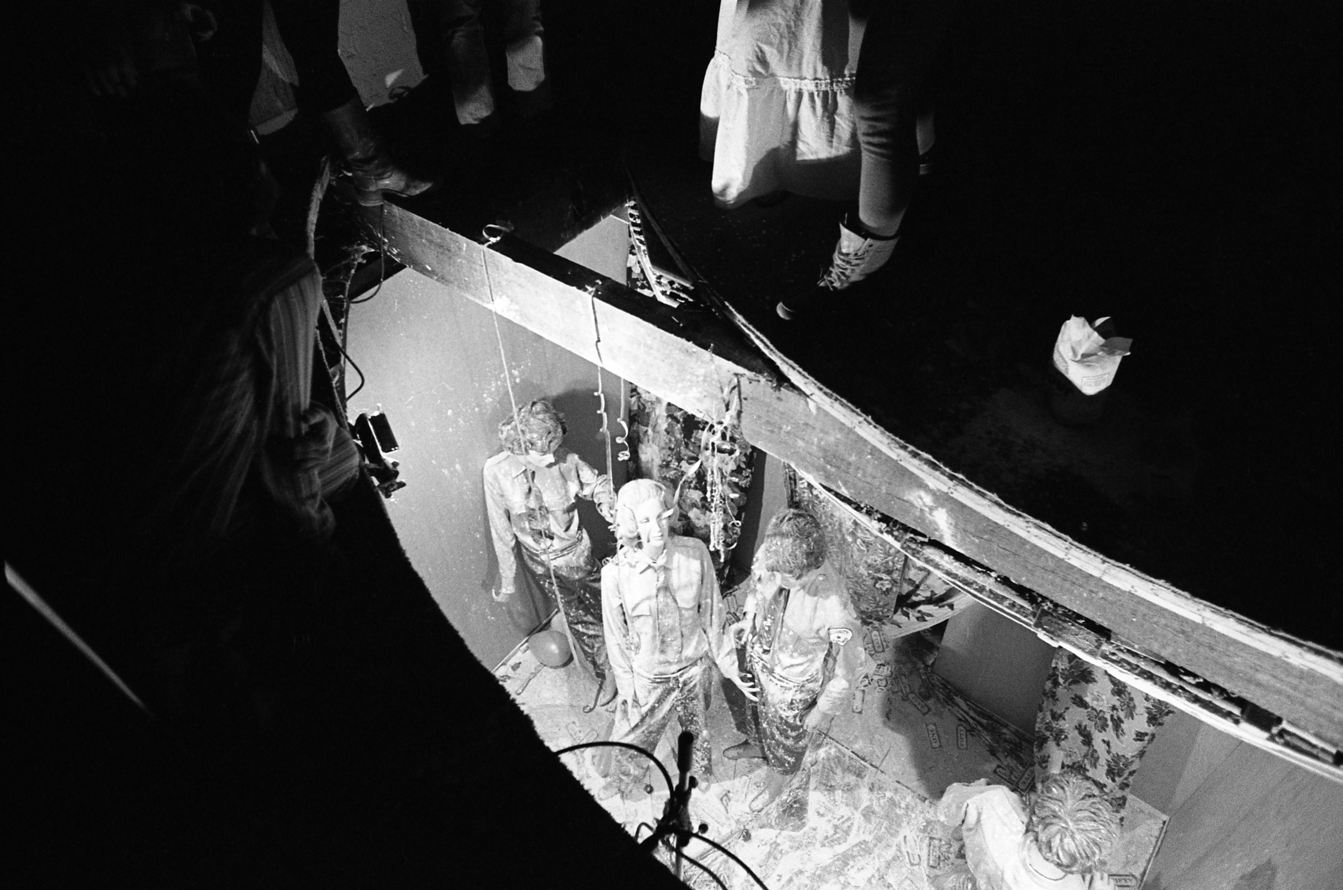 A group of people look up through a cut in a floor. The feet of several others viewing the group can be seen around the cut.