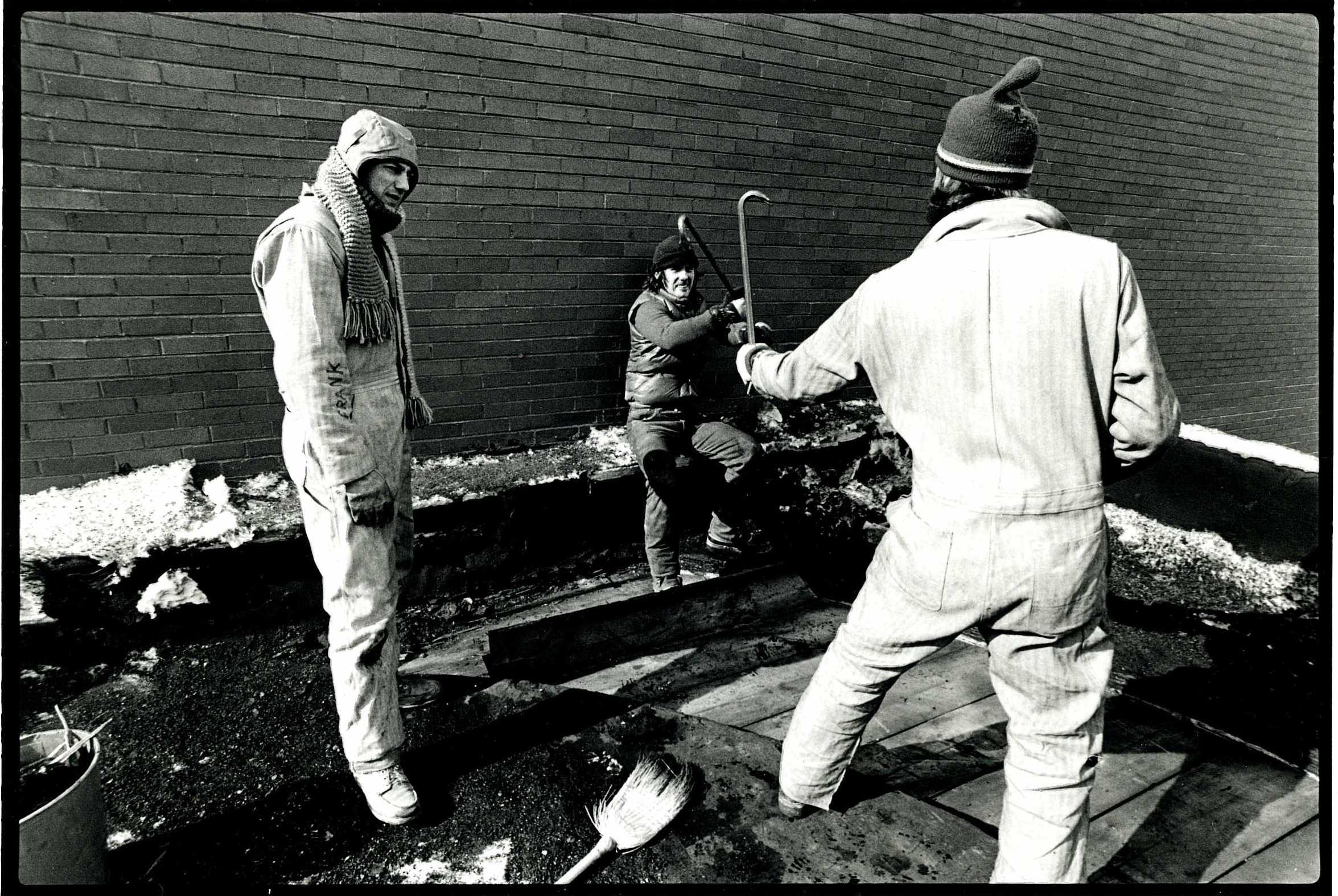 Two people in coveralls pose with crowbars as if ready to battle while another person looks on.