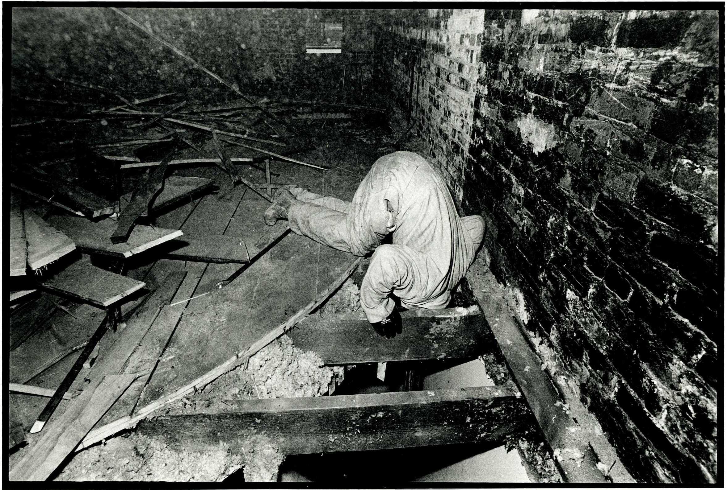 A person in coveralls, supporting themself with the rafters, tilts their whole body down into a hole in a floor.