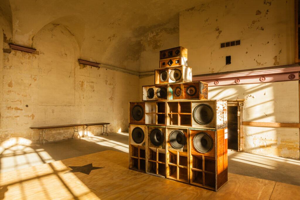 A sculpture of stacked speakers in ascending size from top to bottom on top of a plywood floor with a black star.