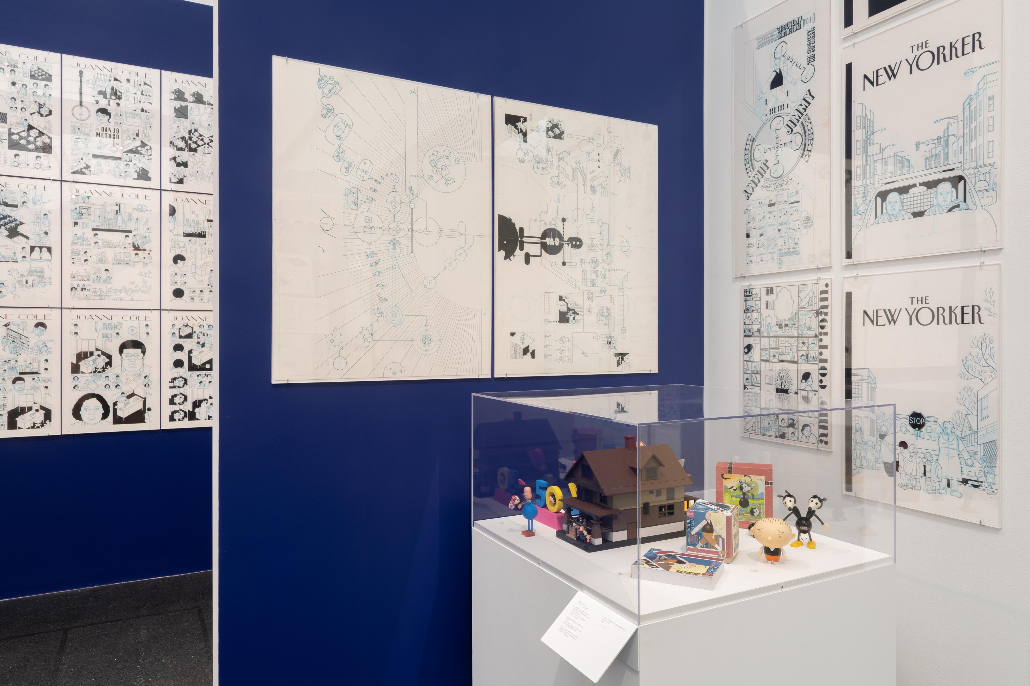 A view of two rooms separated by a blue divider. On the walls are several different comics and drawings, including two New Yorker covers. On a podium in the foreground is several small 3D models of different things.