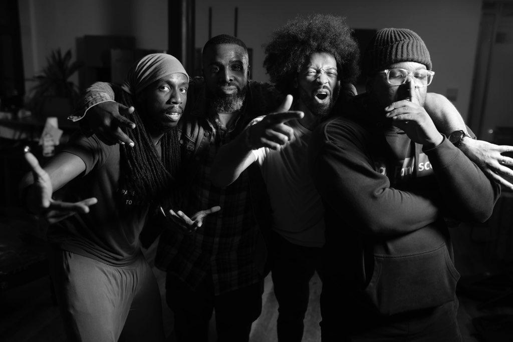 Black-and-white photo of four men posed with arms around each others' shoulders. The man closest to the camera holds his left hand in front of his mouth as if beatboxing.