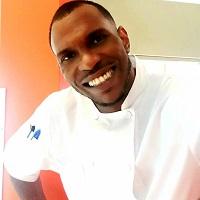 A dark-skinned Black man wearing a white chef coat smiles broadly at the camera.